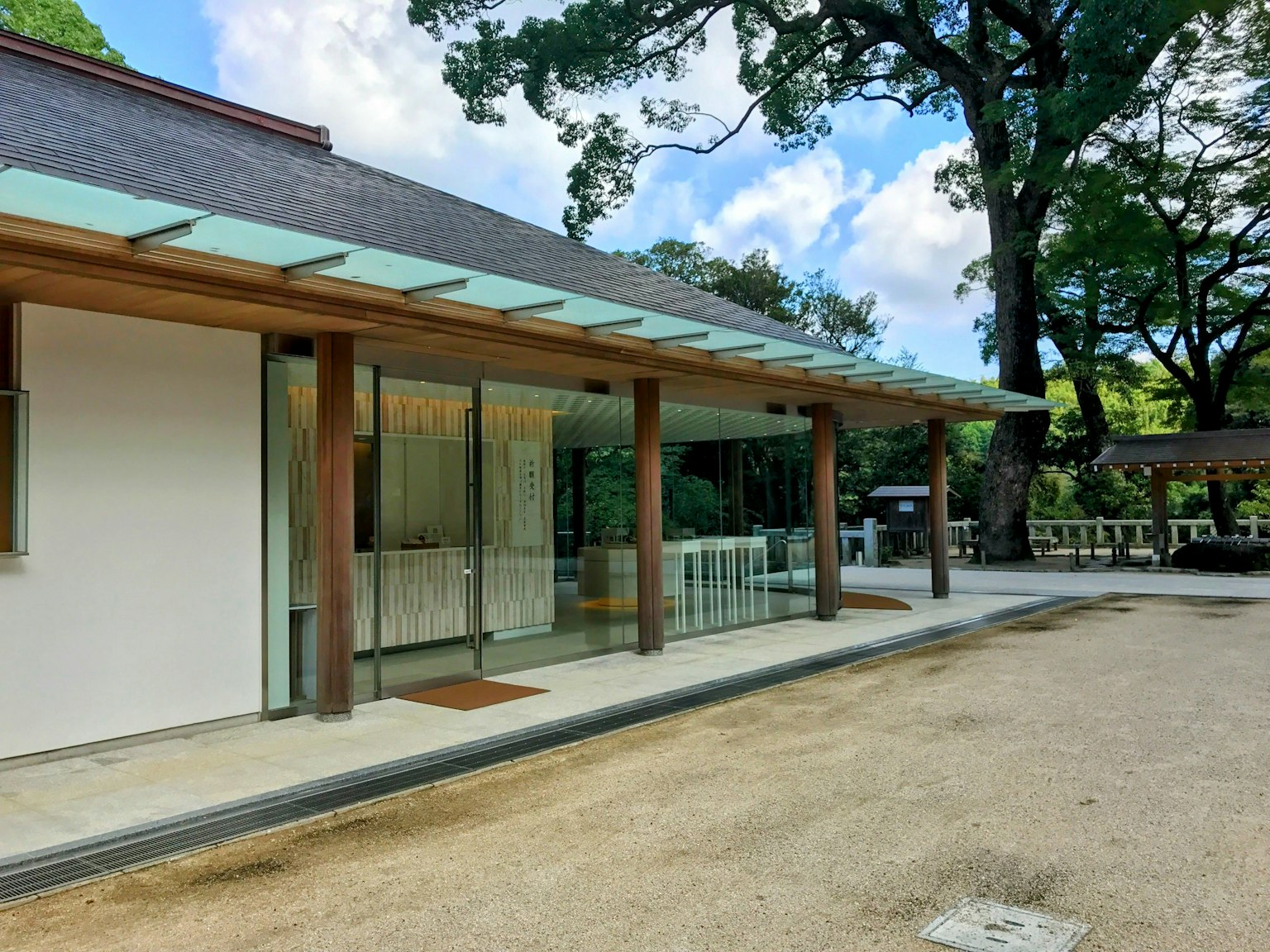 Esterno architettonico moderno con colonne in legno e pareti in vetro sotto un cielo blu e alberi verdi sullo sfondo