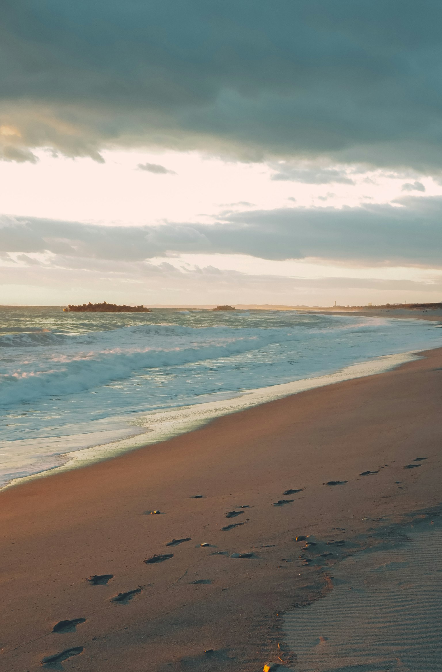 Pemandangan pantai yang tenang dengan ombak lembut dan jejak kaki saat matahari terbenam