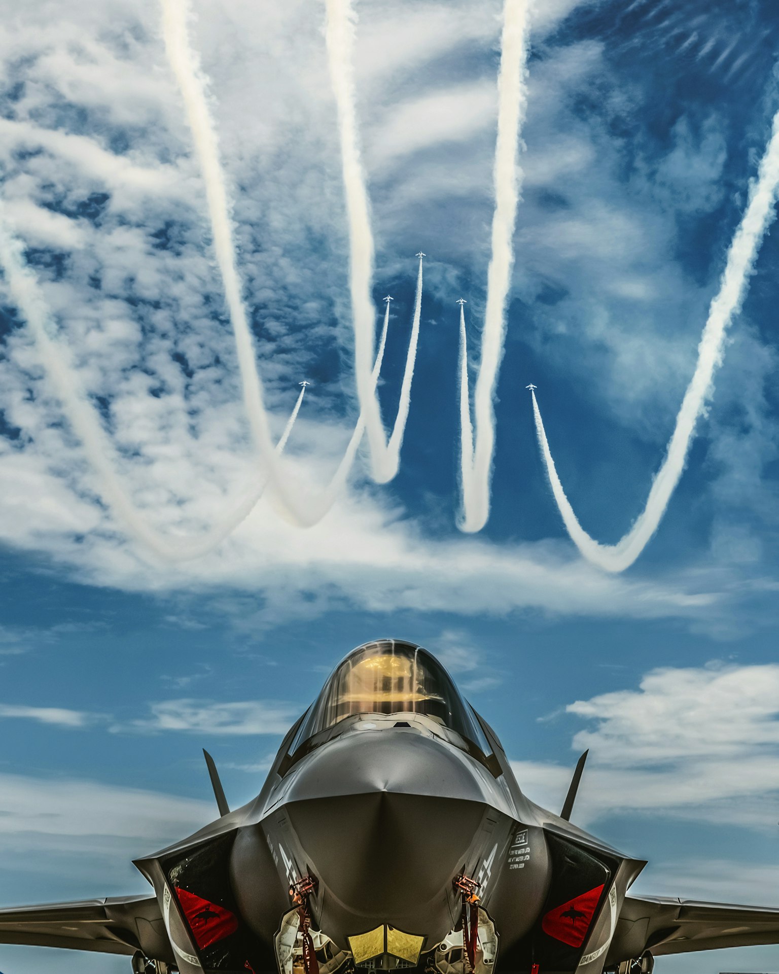 Kampfjet mit Piloten davor und weißen Rauchspuren im blauen Himmel