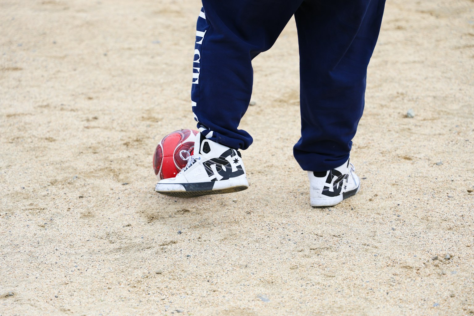 Pieds d'un enfant frappant un ballon de football dans une zone sablonneuse portant des baskets blanches et un pantalon bleu