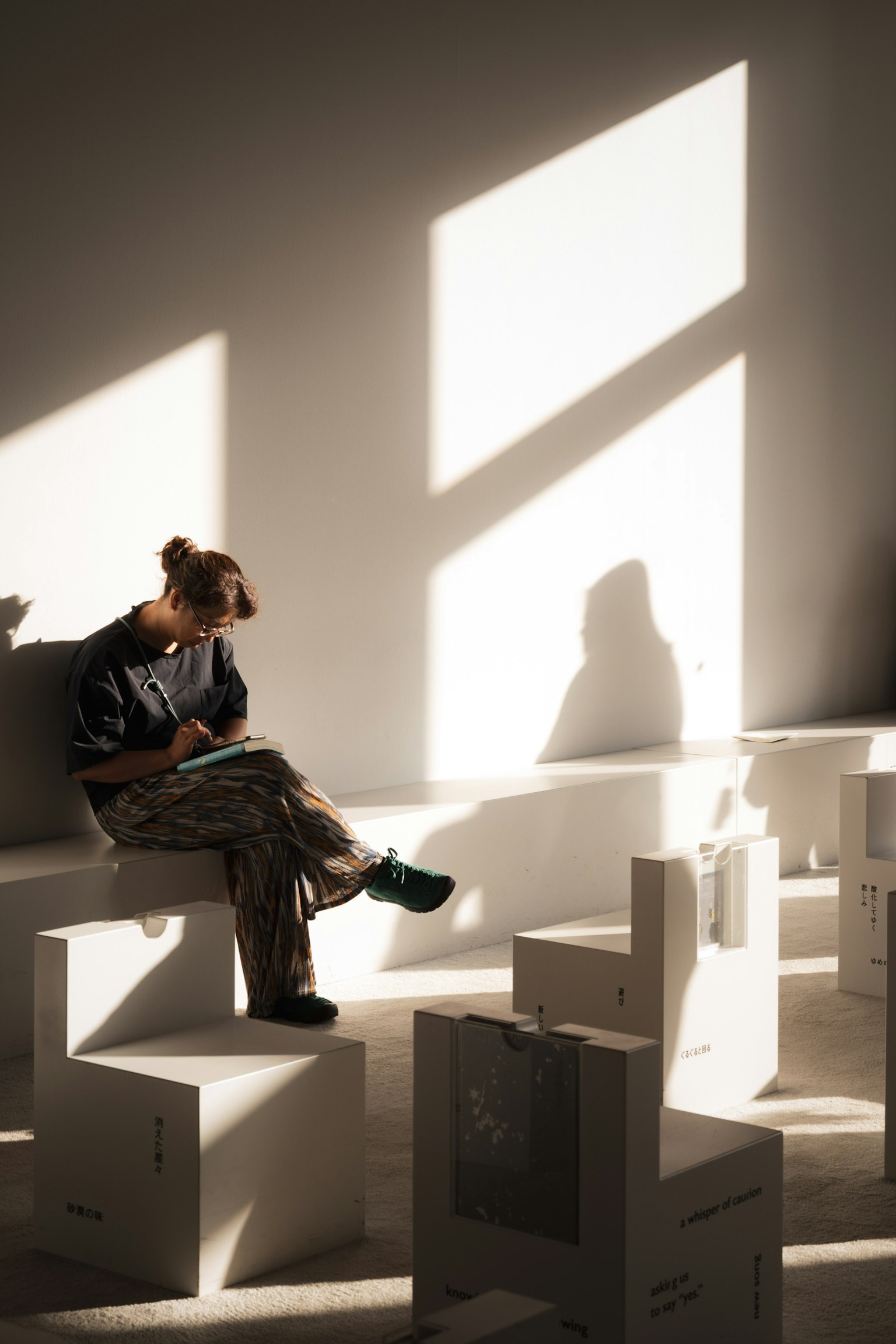女性が白い箱の上に座り、光が差し込む部屋で本を読んでいる