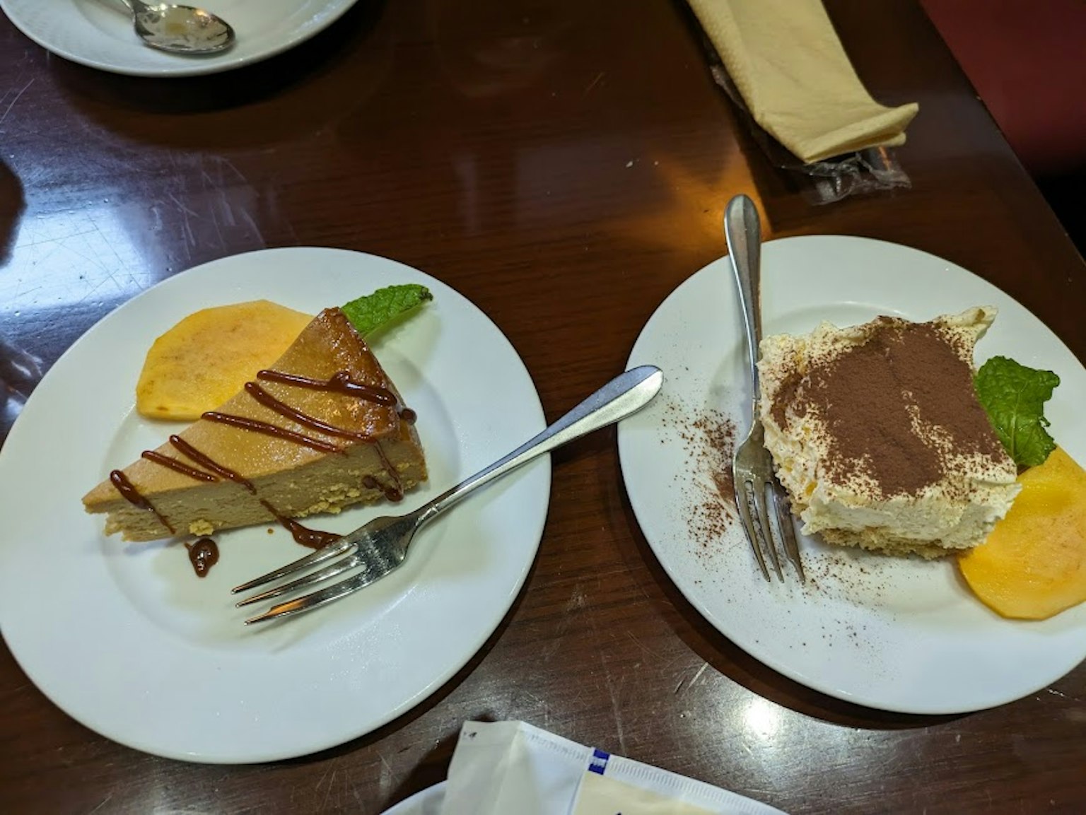 Due piatti di dessert su un tavolo Uno presenta una torta guarnita di caramello con una fetta d'arancia e foglie di menta L'altro è un tiramisù decorato con cacao in polvere e foglie di menta