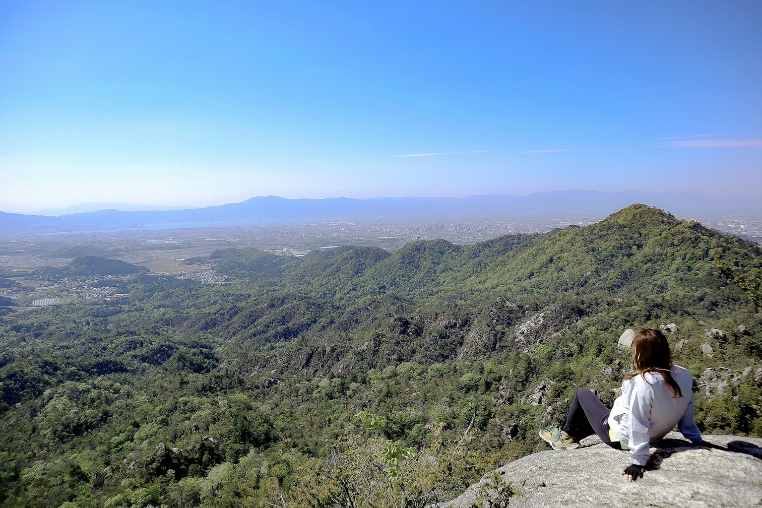 Woman sitting on a mountain peak overlooking a vast green valley and blue sky