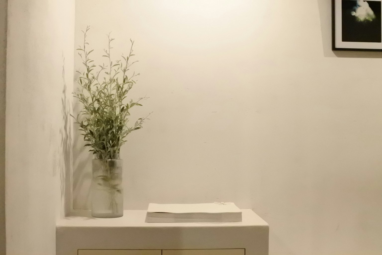 A simple white vase with greenery on a white table against a white wall