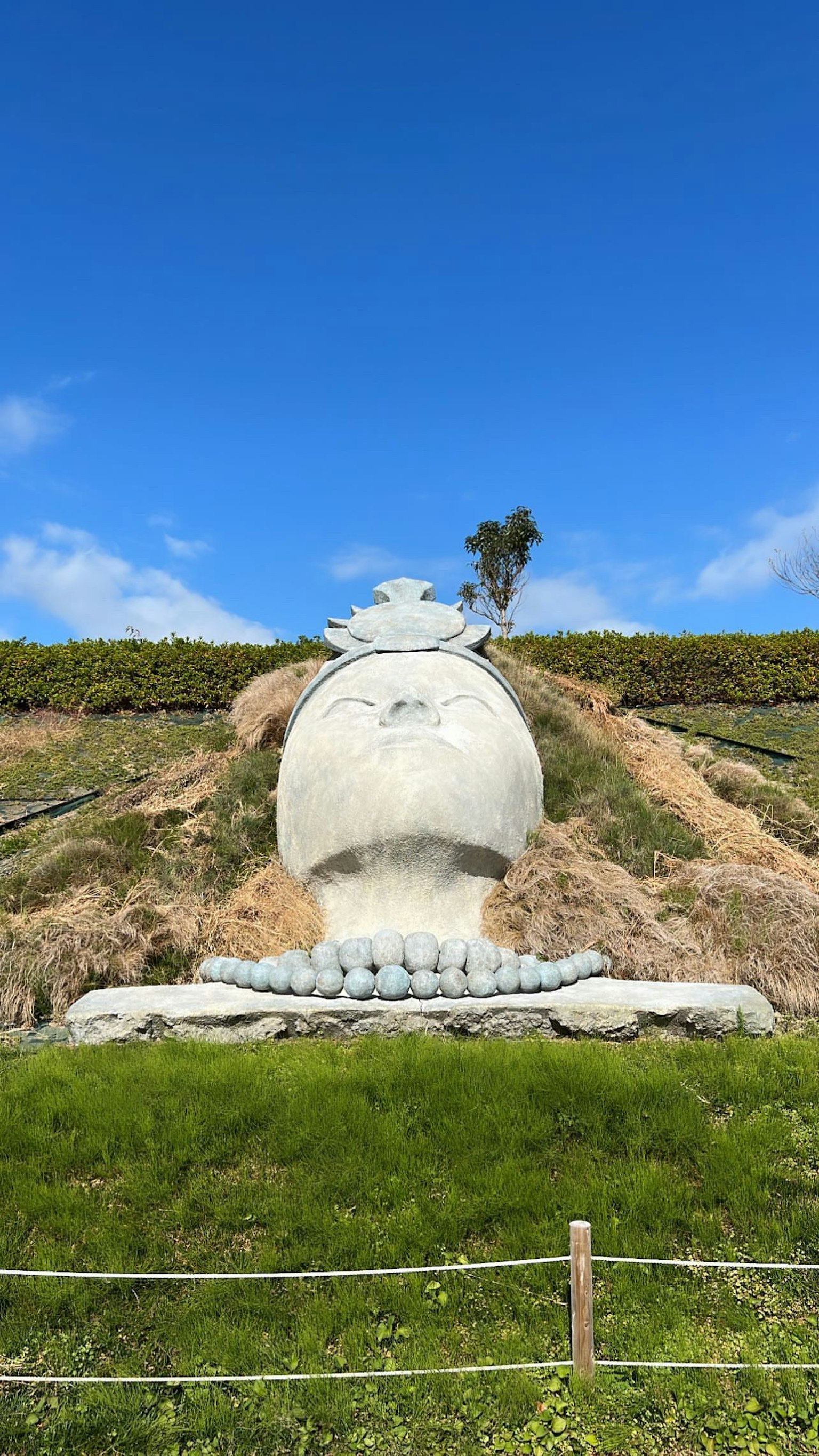 Große Skulptur unter einem blauen Himmel umgeben von grünem Gras und Hügeln mit einem Baum in der Ferne