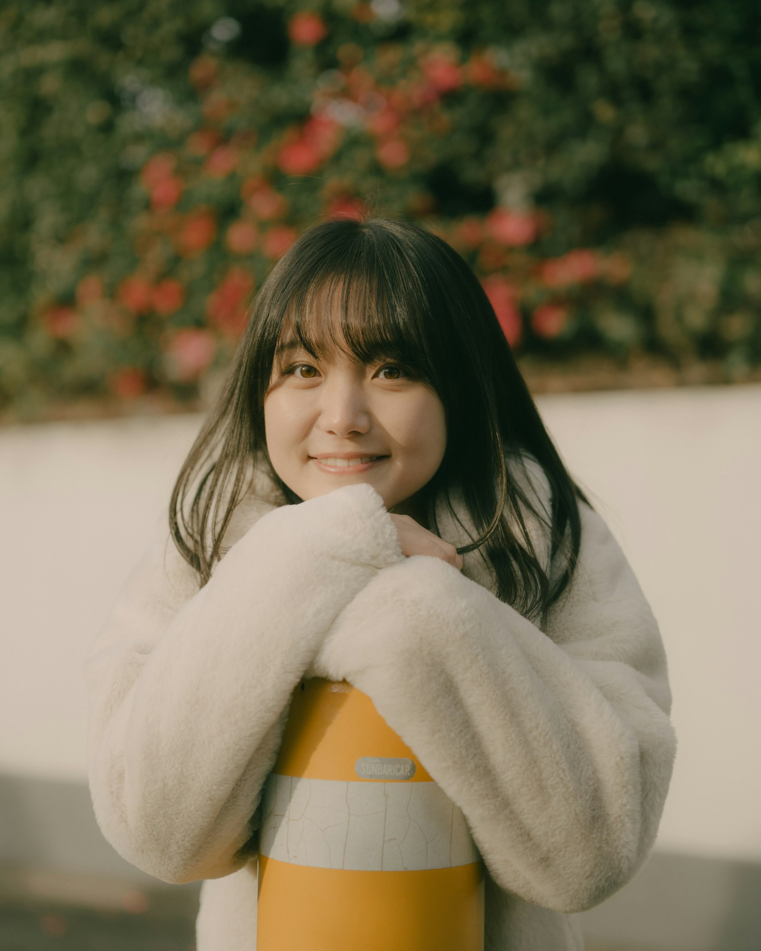 A woman in a white coat smiling while hugging a yellow and white pole with flowers in the background