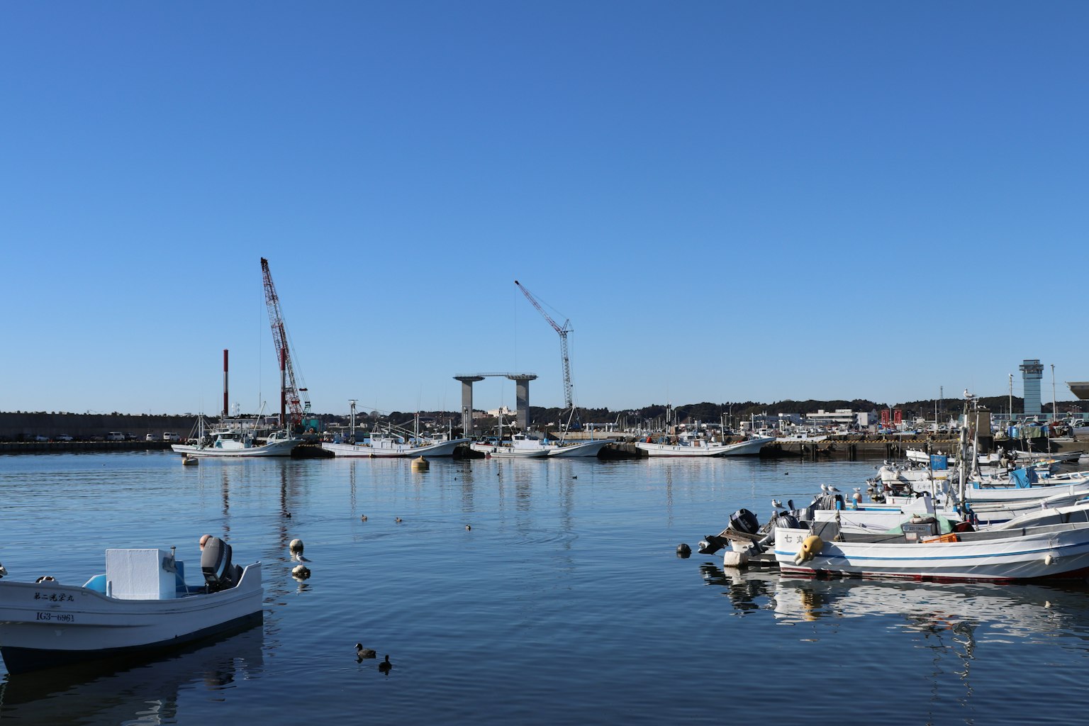 Hafenszene mit Fischerbooten und Baukränen unter einem klaren blauen Himmel