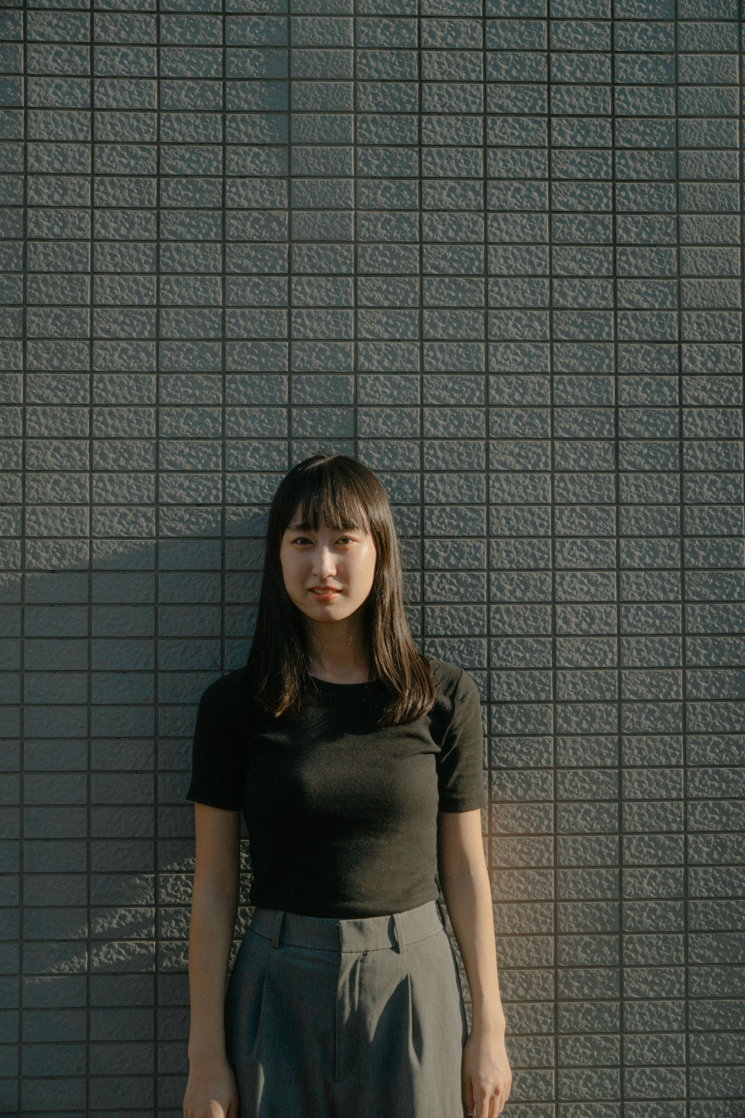 A woman wearing a black shirt and gray pants stands against a gray wall