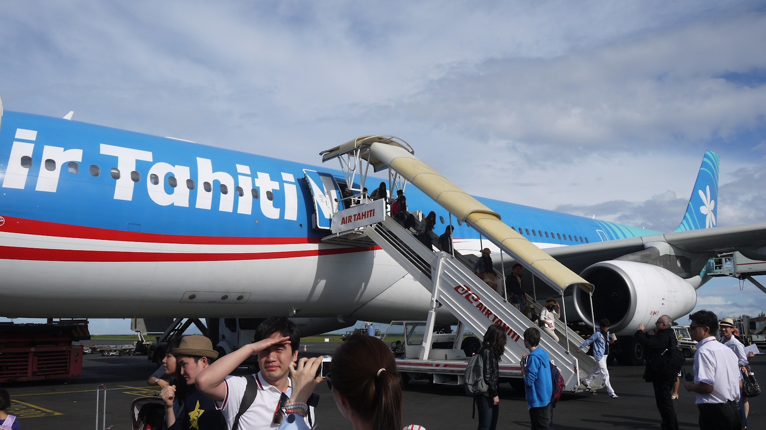 Des passagers descendent de l'avion Air Tahiti Nui avec un ciel bleu et des nuages blancs en arrière-plan