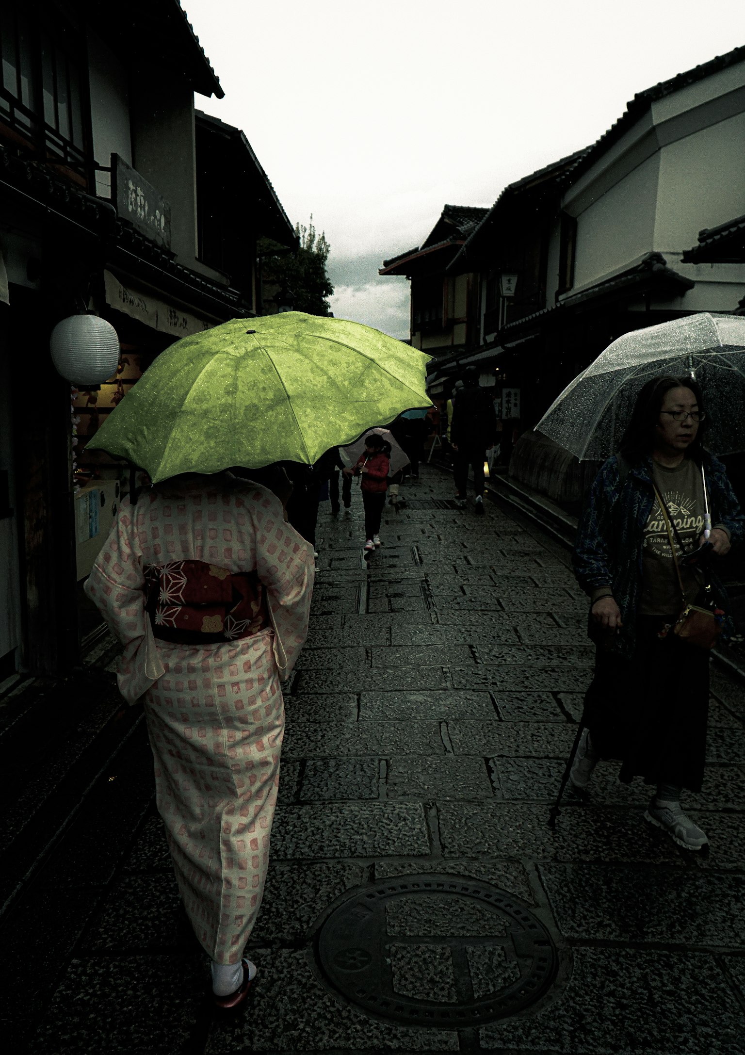 Frau im Kimono, die unter einem grünen Regenschirm in einer regnerischen Straße geht