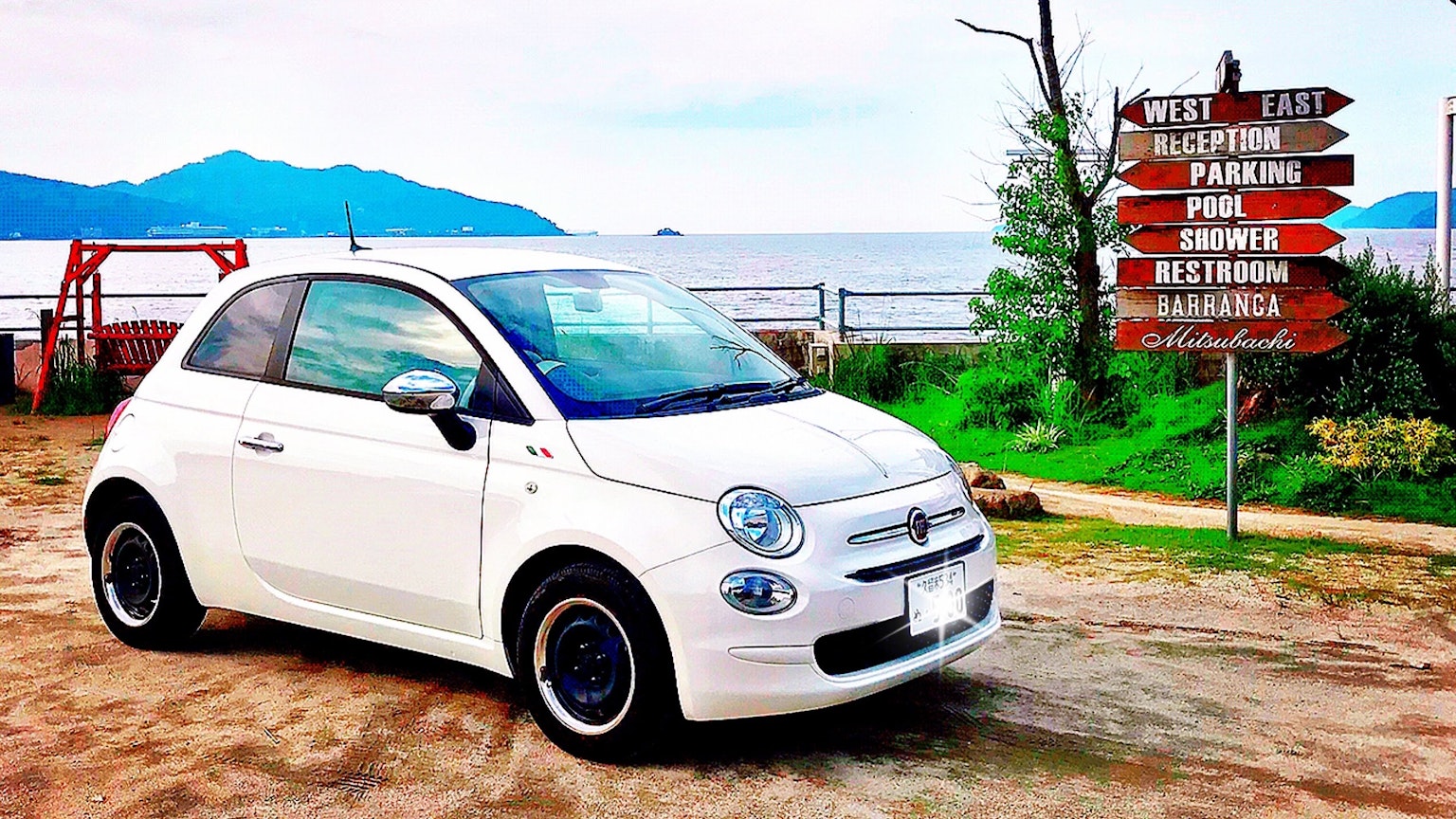 Một chiếc Fiat 500 màu trắng đậu gần bãi biển với biển chỉ dẫn ở phía sau