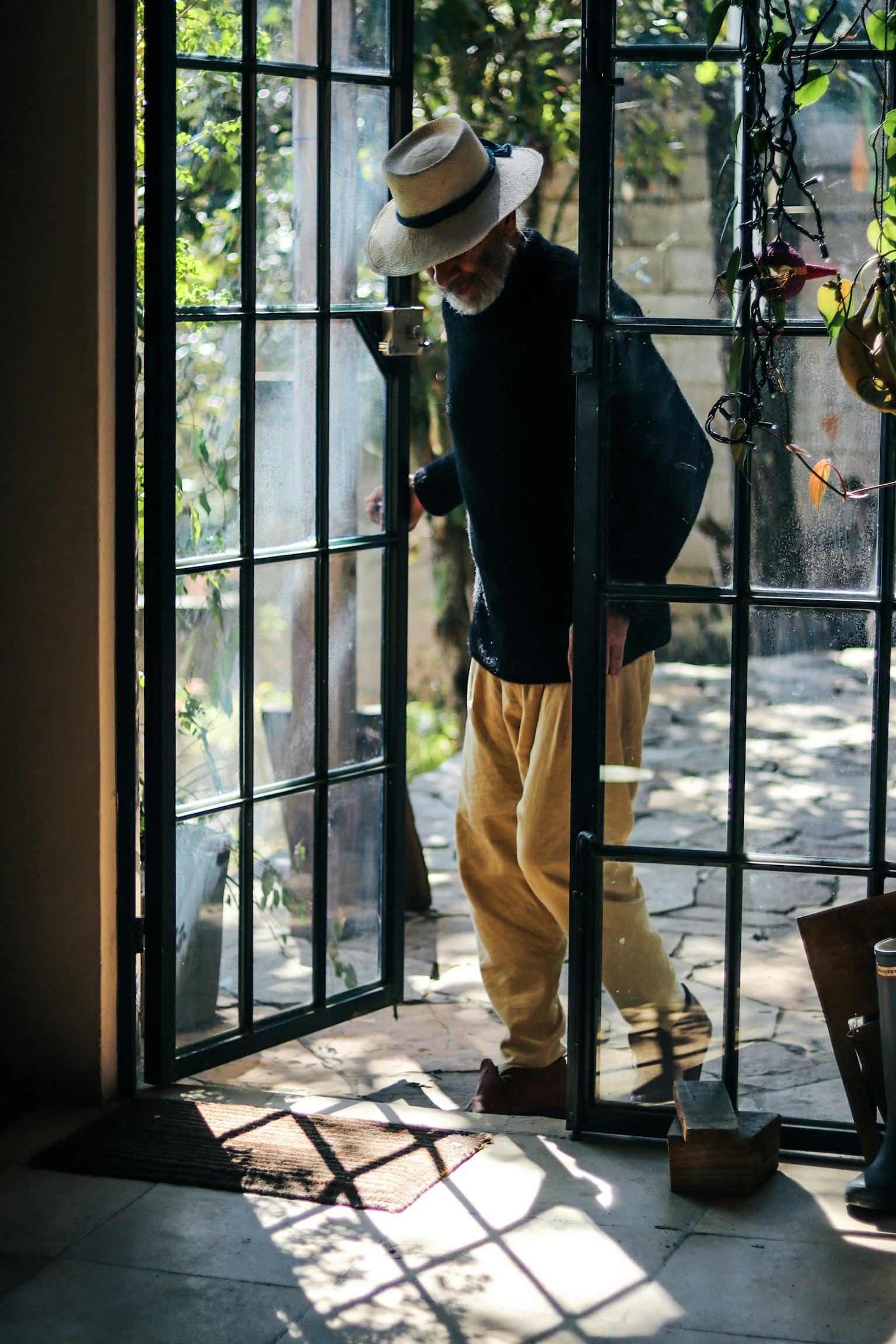 Man stepping through an open door wearing a hat with plants and sunlight visible