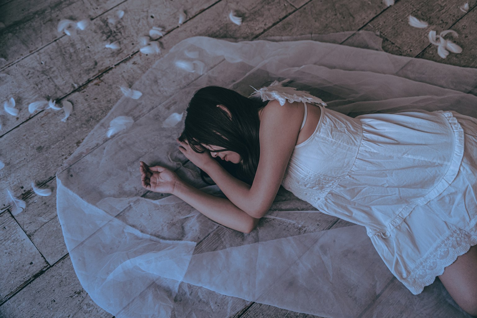 白いドレスを着た女性が床に横たわり、薄い布に包まれている周囲には羽毛が散らばっている