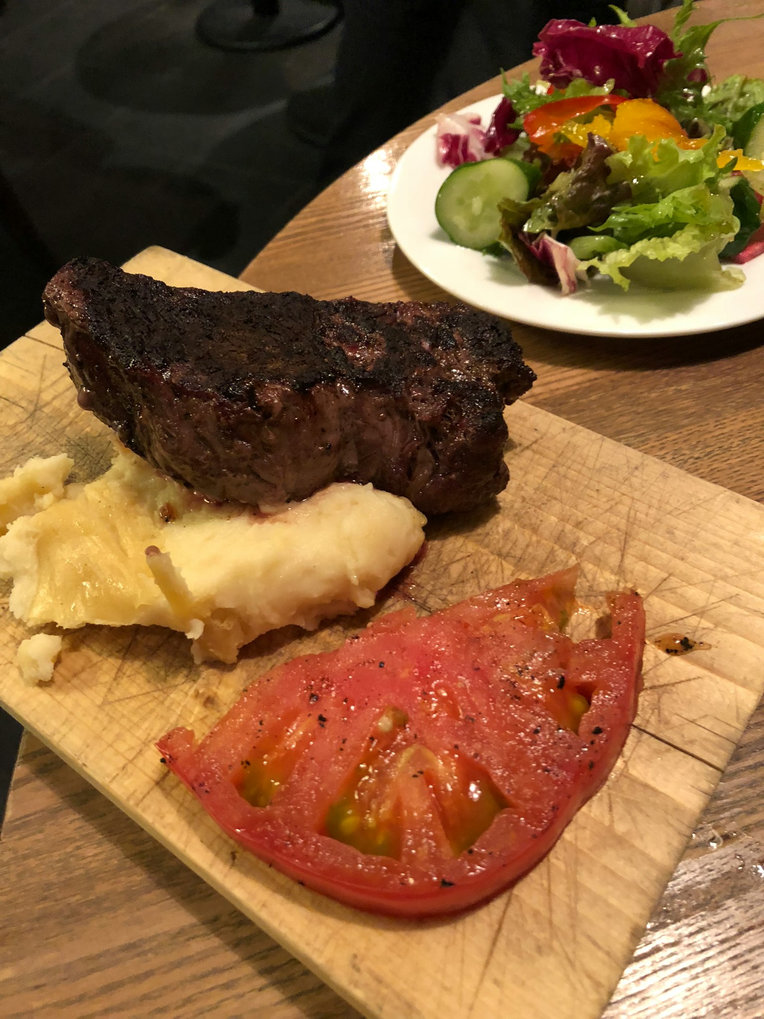 A grilled steak with mashed potatoes and a slice of tomato on a wooden plate
