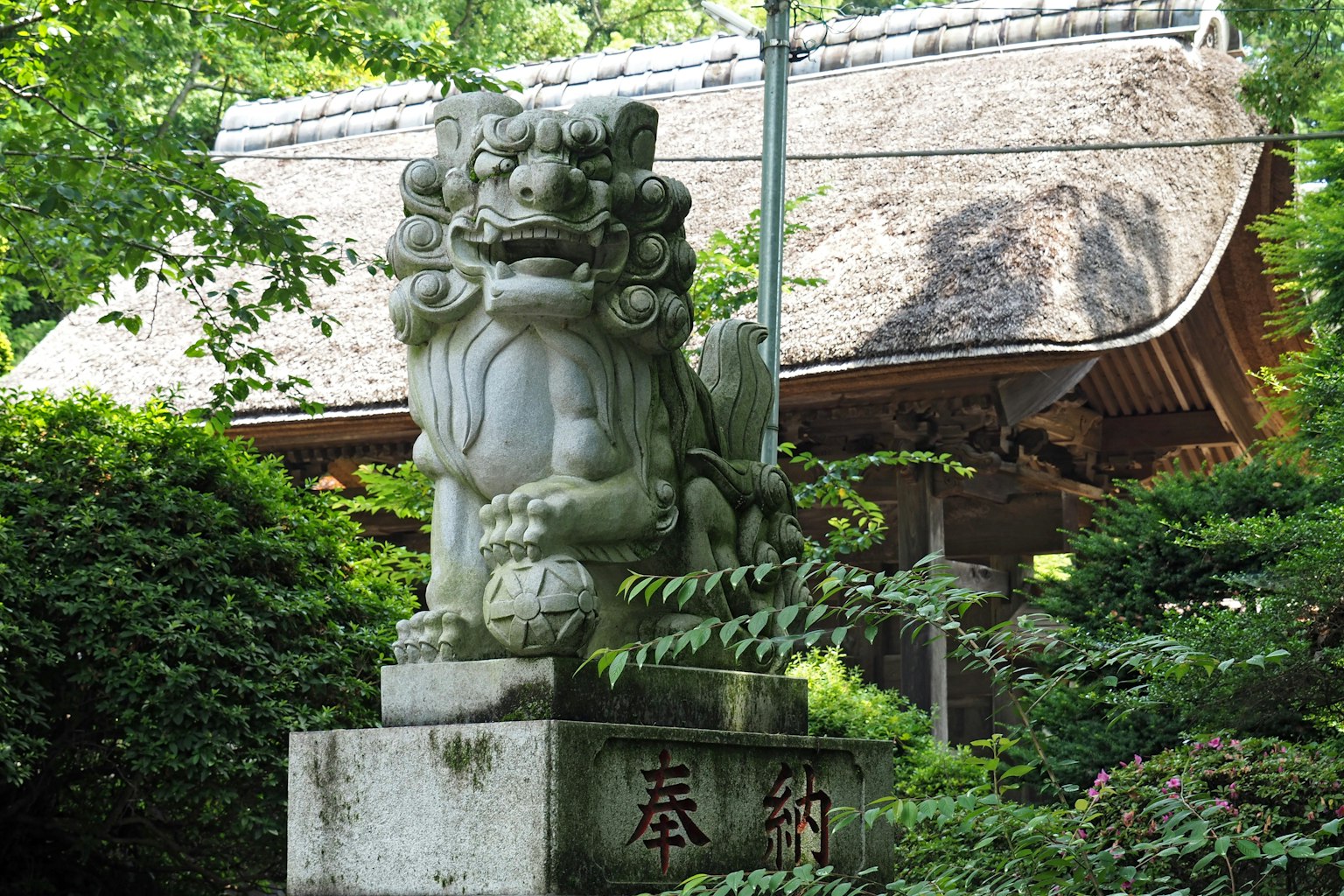 Stone guardian lion statue amidst lush greenery with a traditional thatched roof building in the background