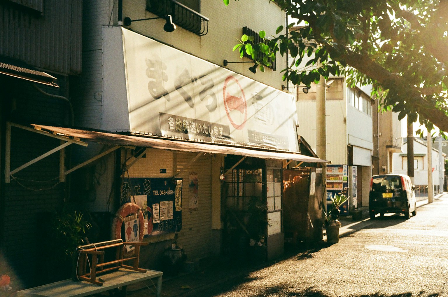 ภายนอกของร้านเล็กที่มีแสงแดดส่องถึง ป้ายและเก้าอี้หันหน้าไปทางถนนที่มีใบไม้เขียวขจีรอบๆ