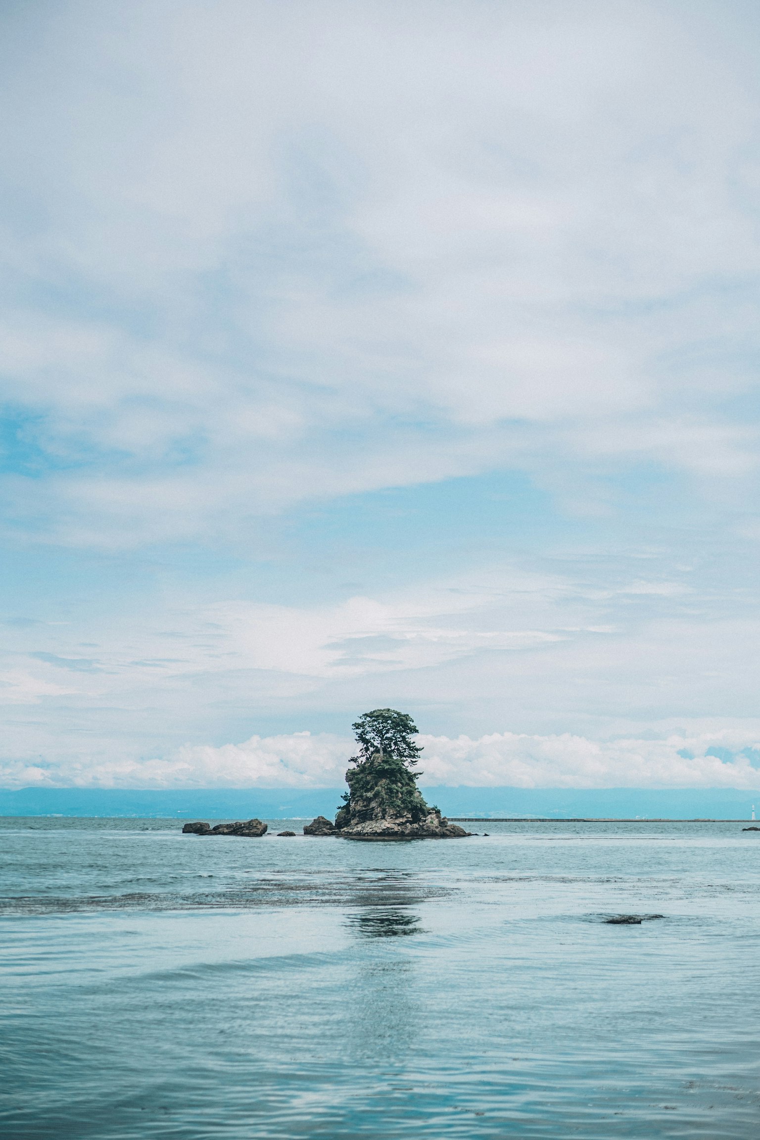 A small island with trees surrounded by calm blue waters and a cloudy sky