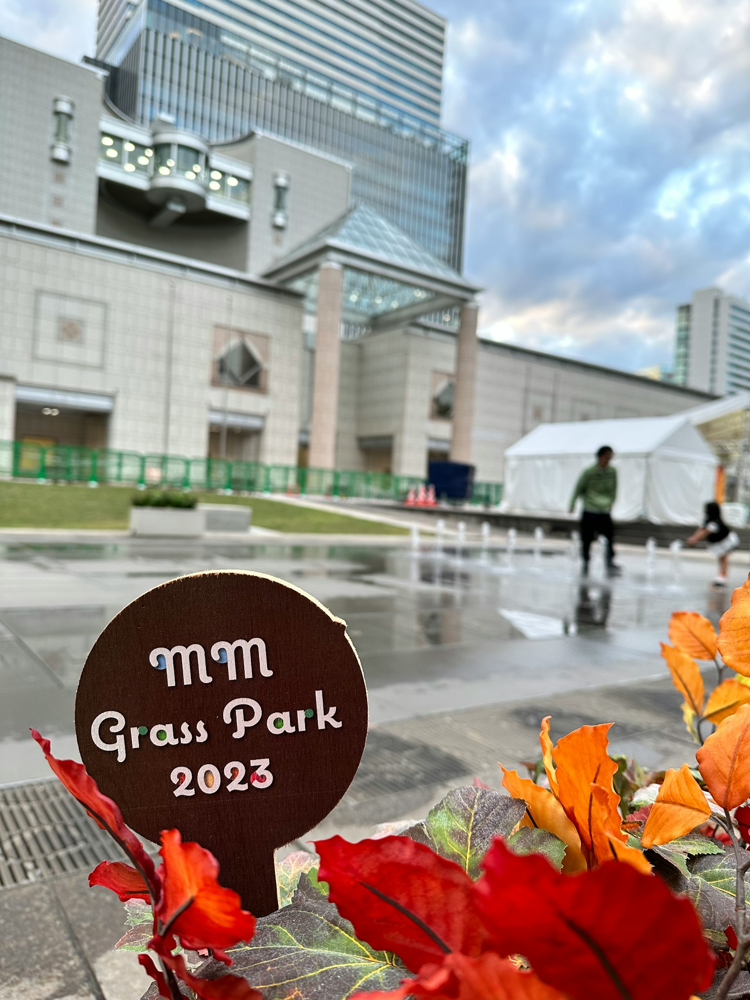 View of MM Grass Park 2023 sign with modern building and autumn leaves in the background