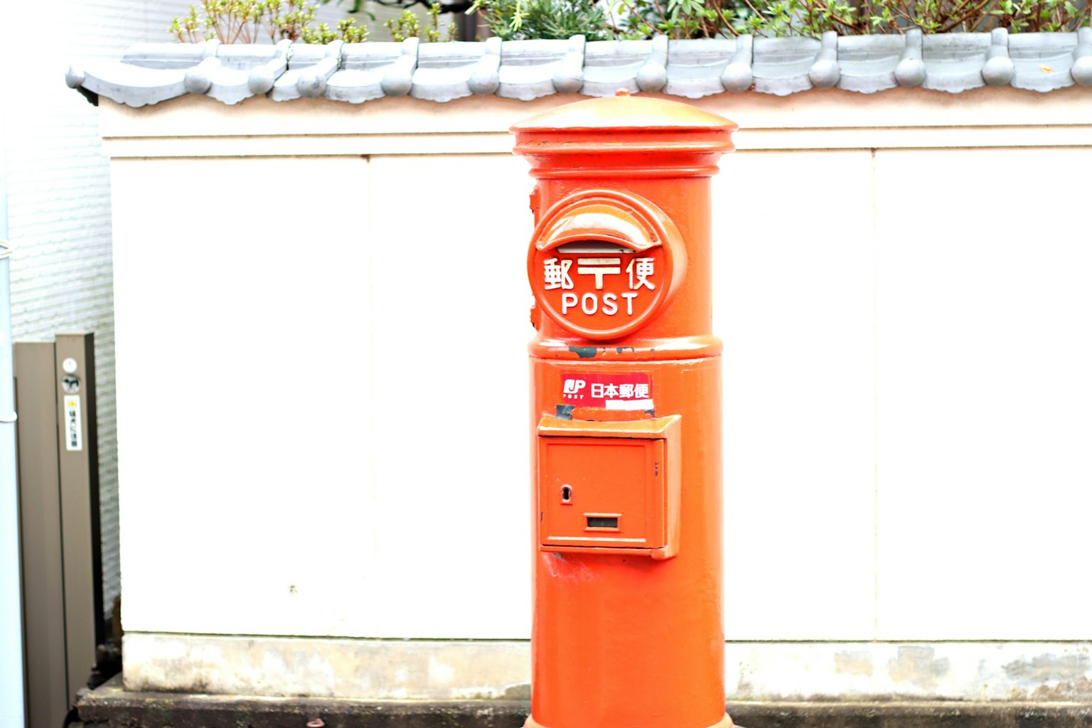 An orange post box stands in front of a white wall