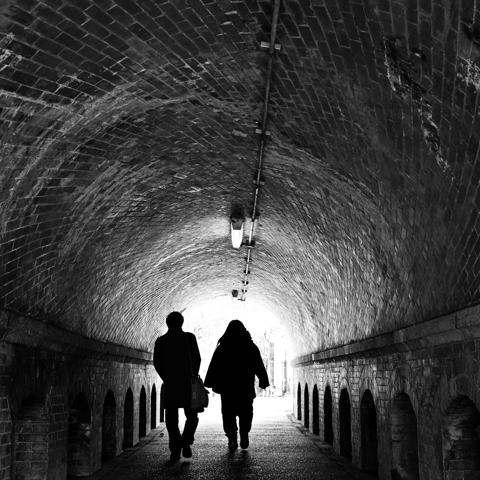 Silhouettes of two people walking through a brick tunnel with bright exit