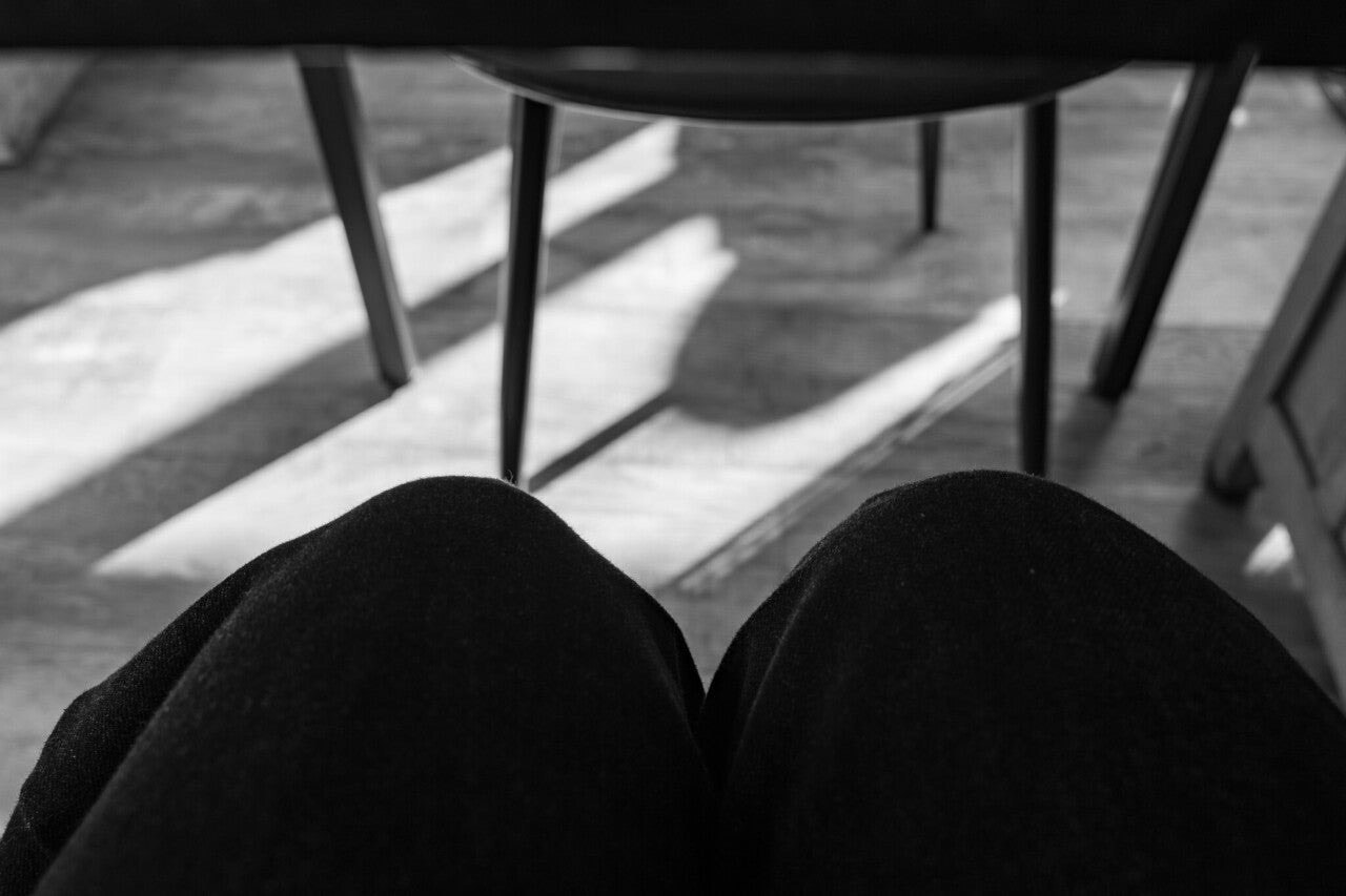 Black and white image showing knees in black pants with chair shadows