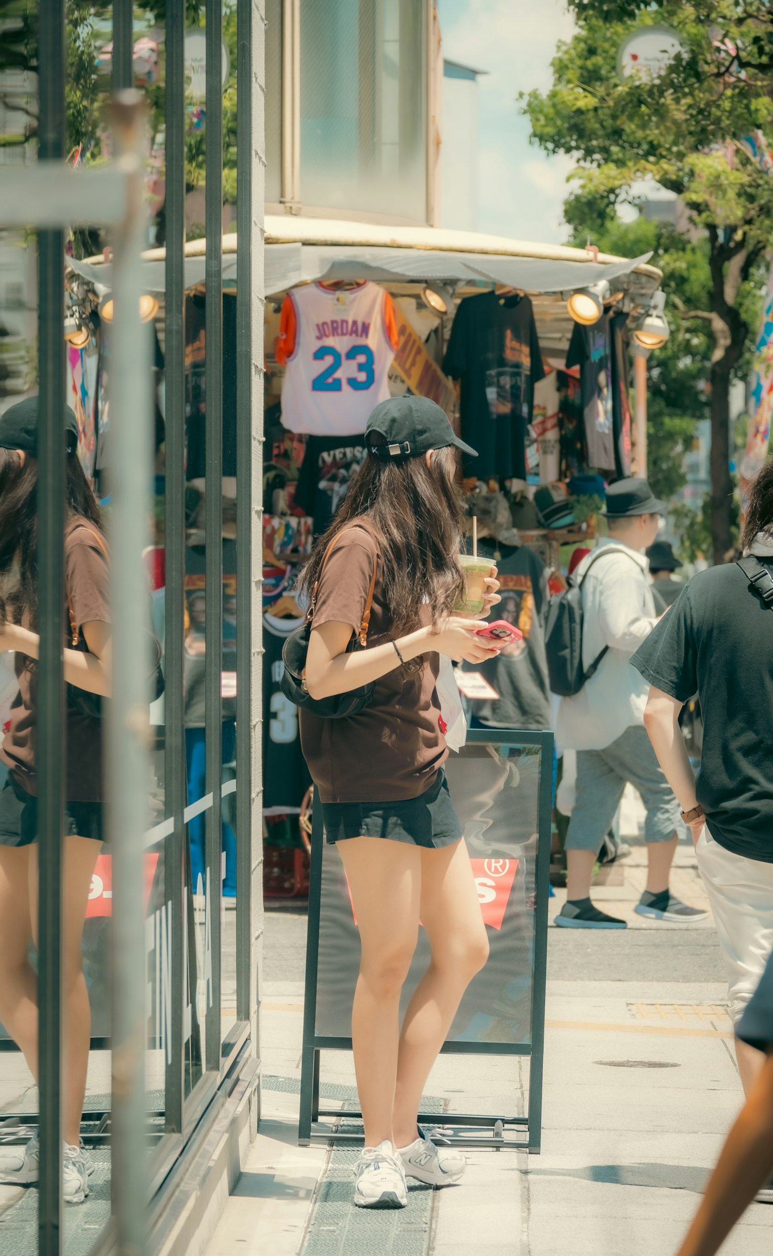 Wanita muda memegang minuman berdiri di depan toko dengan kaos ditampilkan di latar belakang