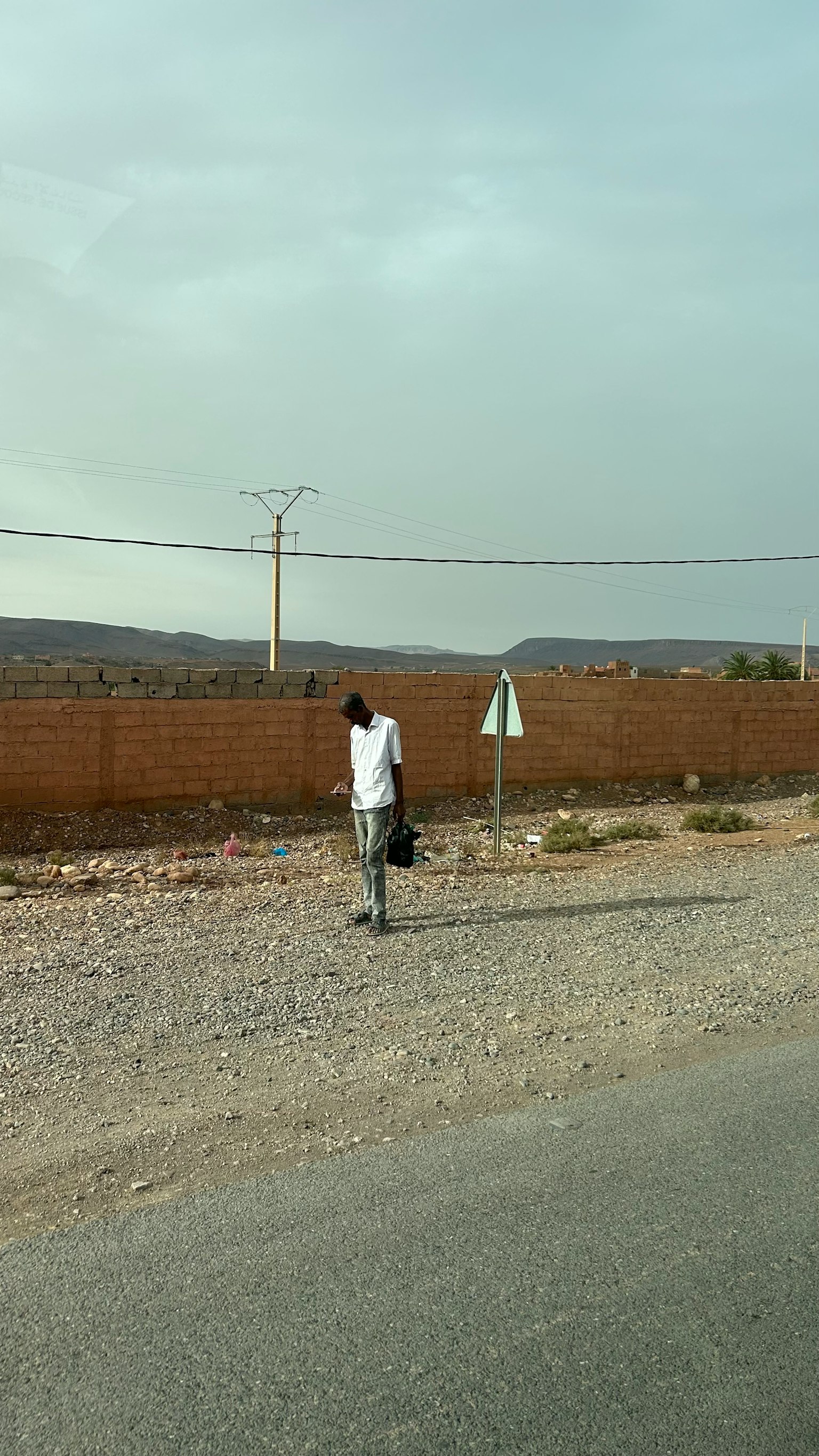 Man walking on a gravel road wearing a white shirt holding a bag with a brown wall in the background