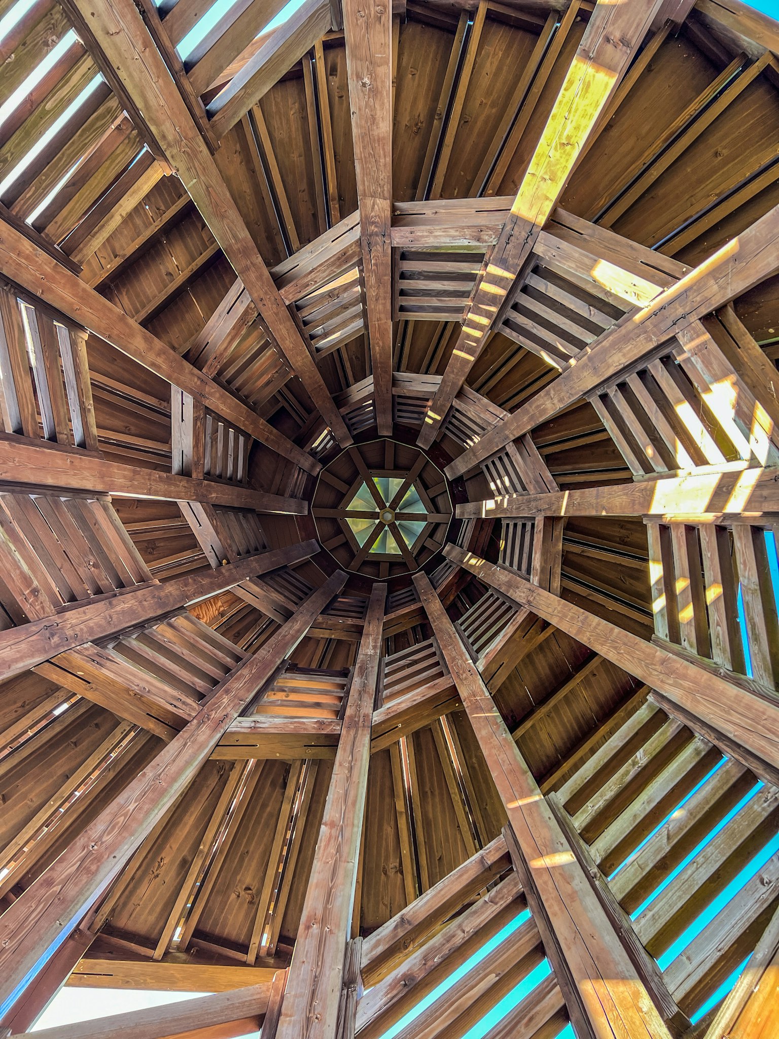 Vue de l'intérieur d'une structure en bois regardant vers le haut montrant des motifs radiaux et un ciel bleu brillant