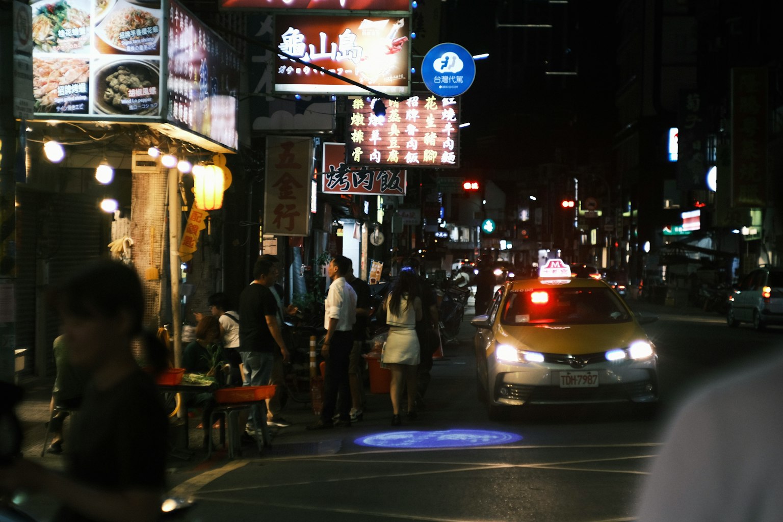 Cảnh đường phố ban đêm với nhà hàng và một chiếc taxi đang chạy qua