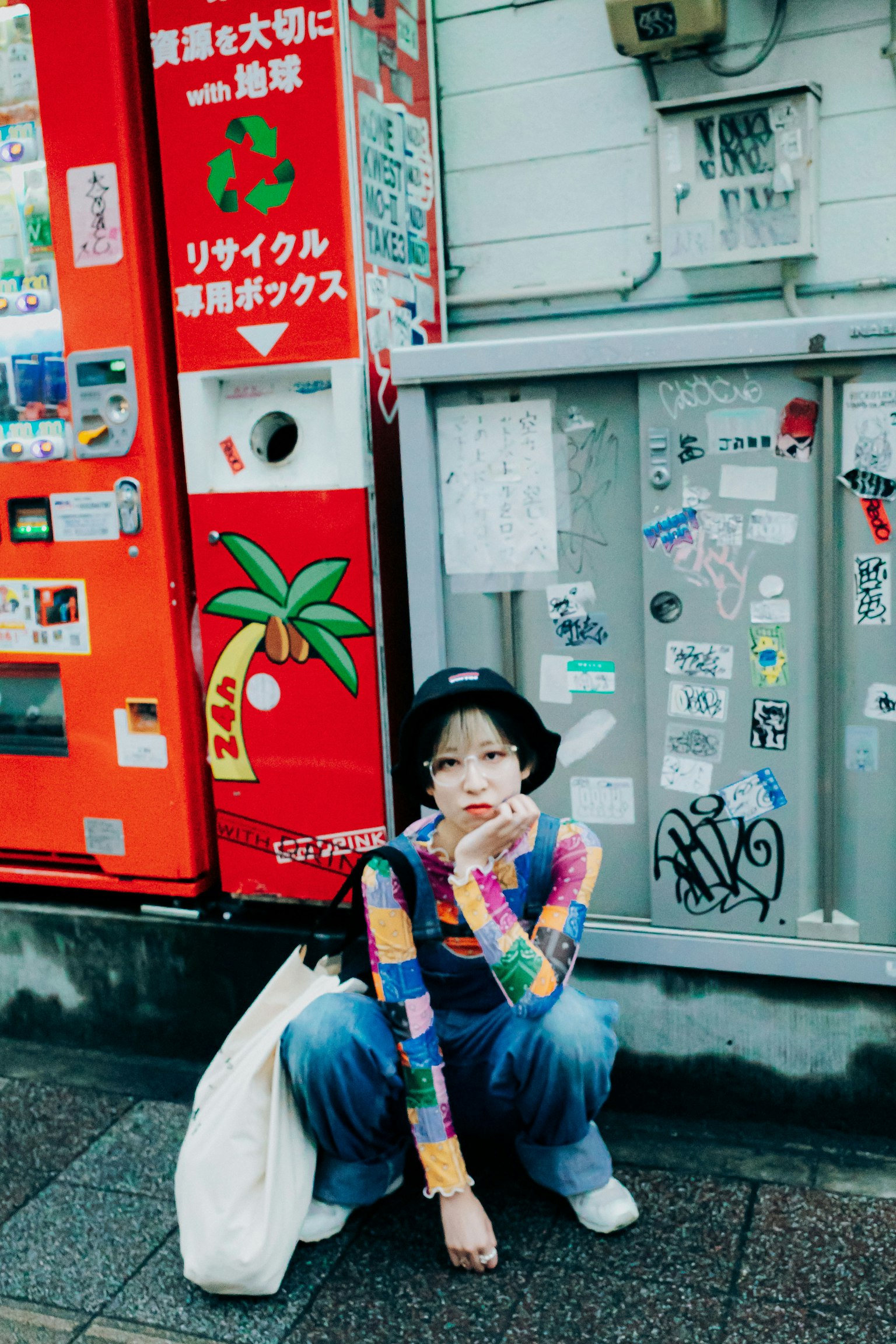 다채로운 상의를 입은 여성이 거리에서 앉아 있으며 배경에는 자판기와 스티커가 있다
