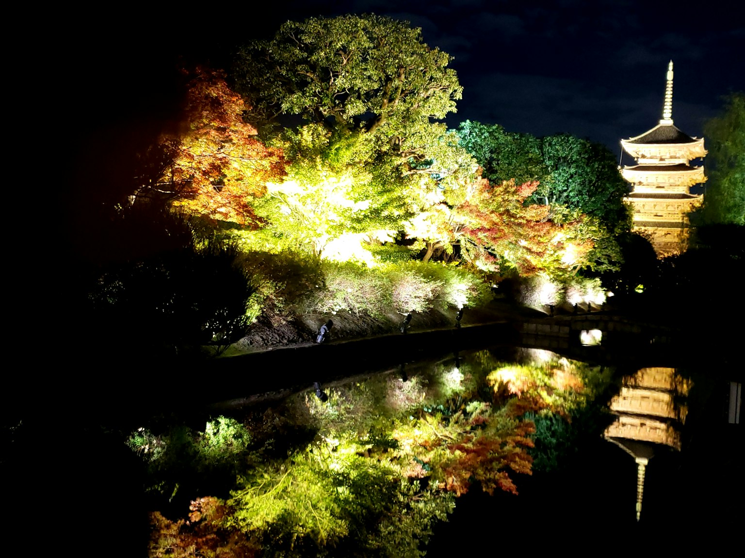 Scena serena di un parco notturno con alberi colorati e una pagoda riflessa nell'acqua