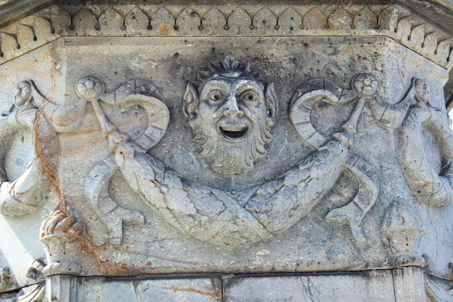 Colonna in pietra scolpita con un volto che presenta occhi grandi e una bocca sorridente circondata da motivi simili a serpenti