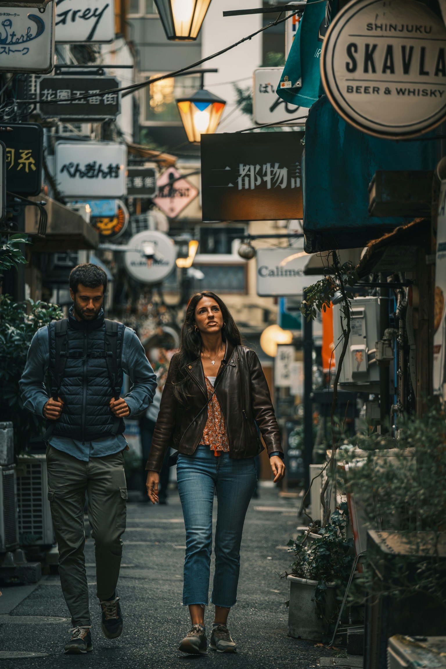 Man and woman walking in a narrow alley surrounded by shop signs and greenery