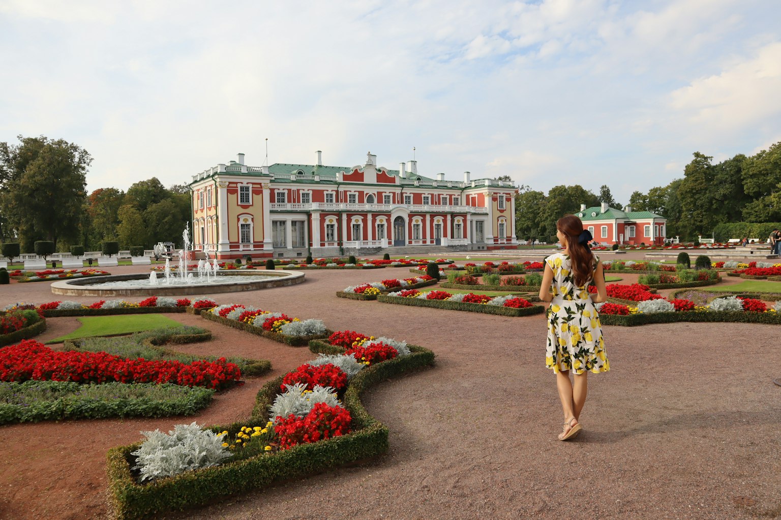 Una donna in abito giallo che cammina in un giardino colorato con un grande edificio sullo sfondo