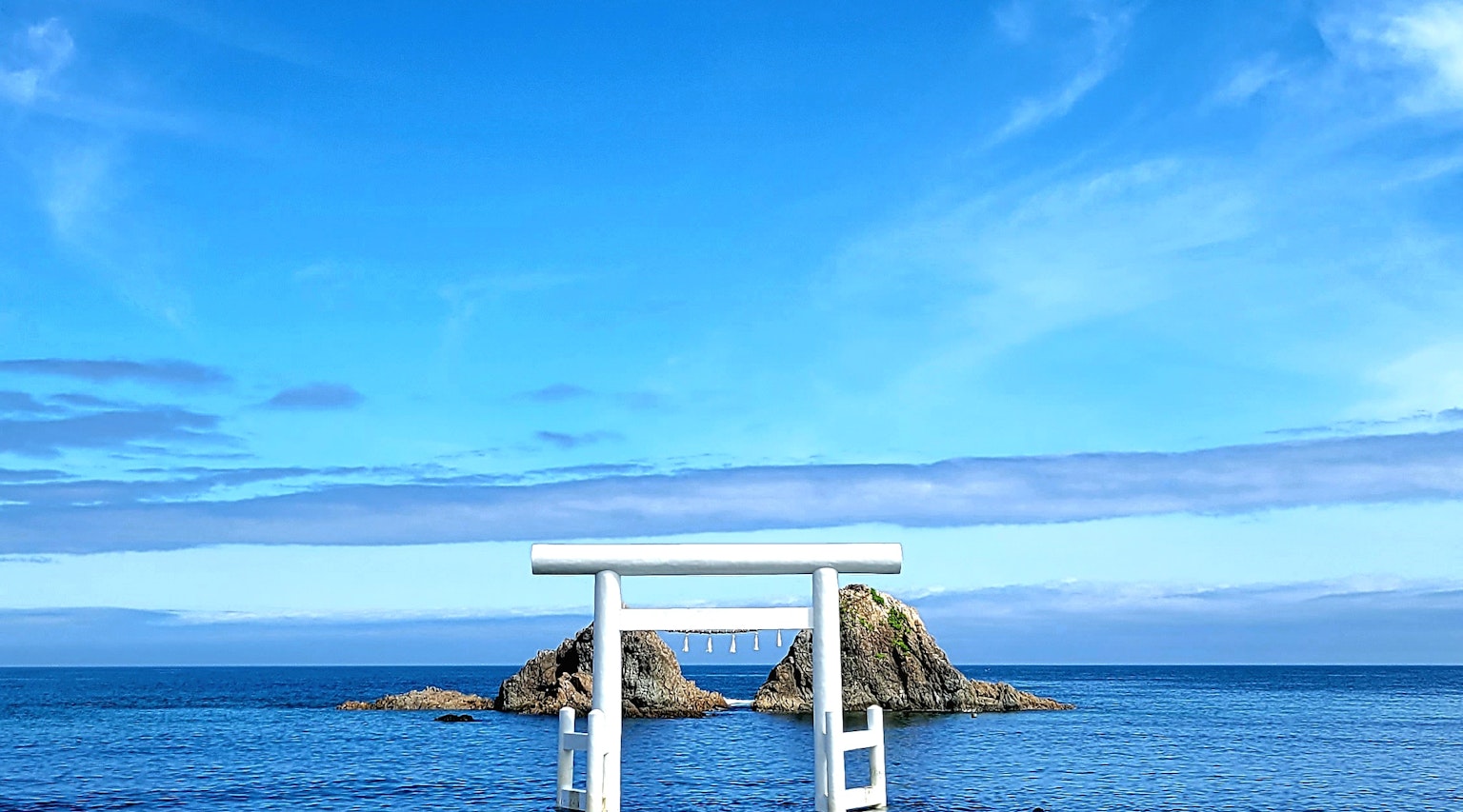 Gerbang torii putih dengan pulau berbatu di bawah langit dan laut biru