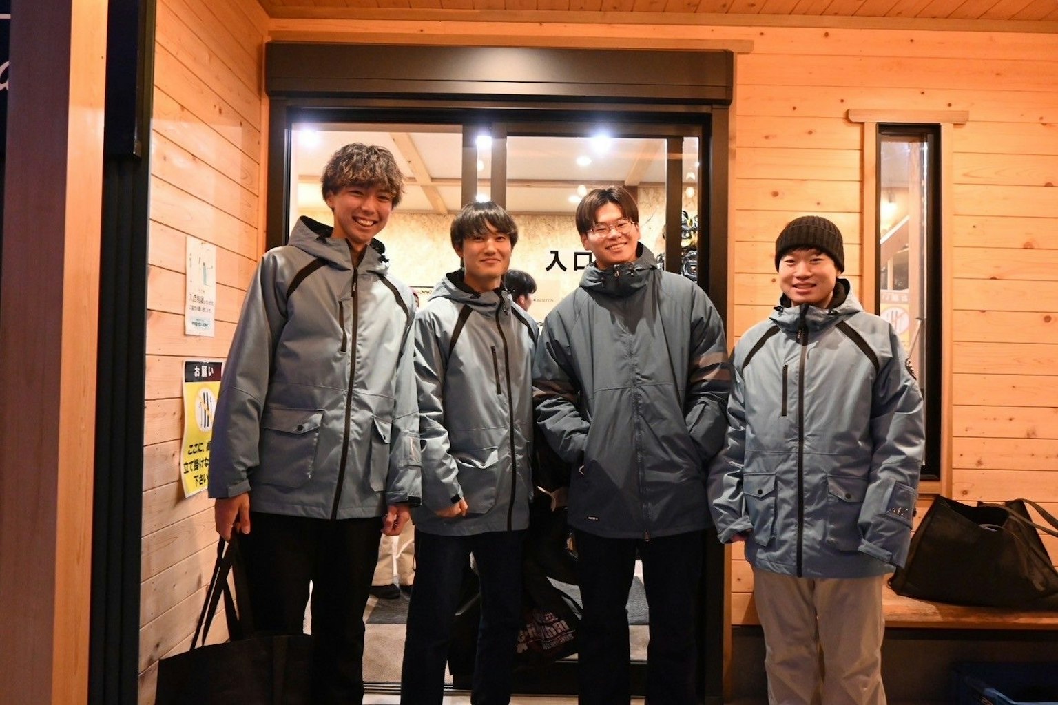 Four men wearing blue jackets posing at the entrance of a wooden building