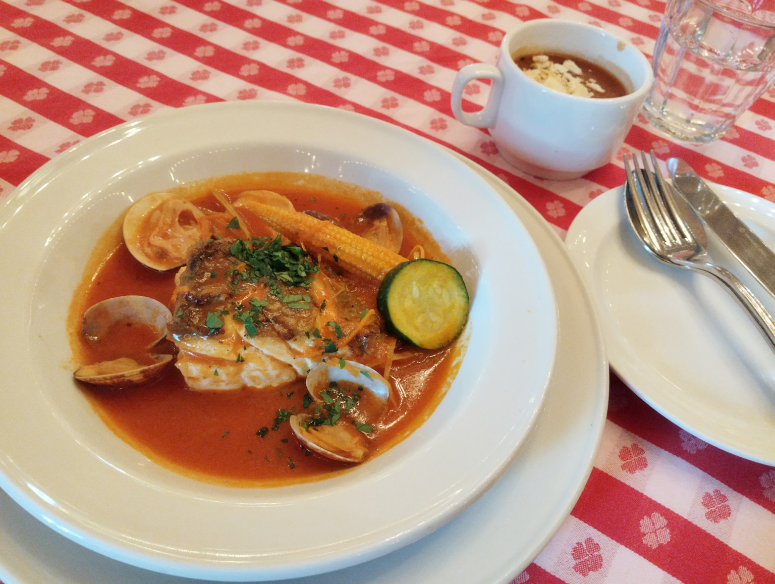 Soupe de fruits de mer servie dans un bol avec des garnitures de citron et de courgette, nappe à carreaux rouges et blancs, café et verre d'eau en arrière-plan