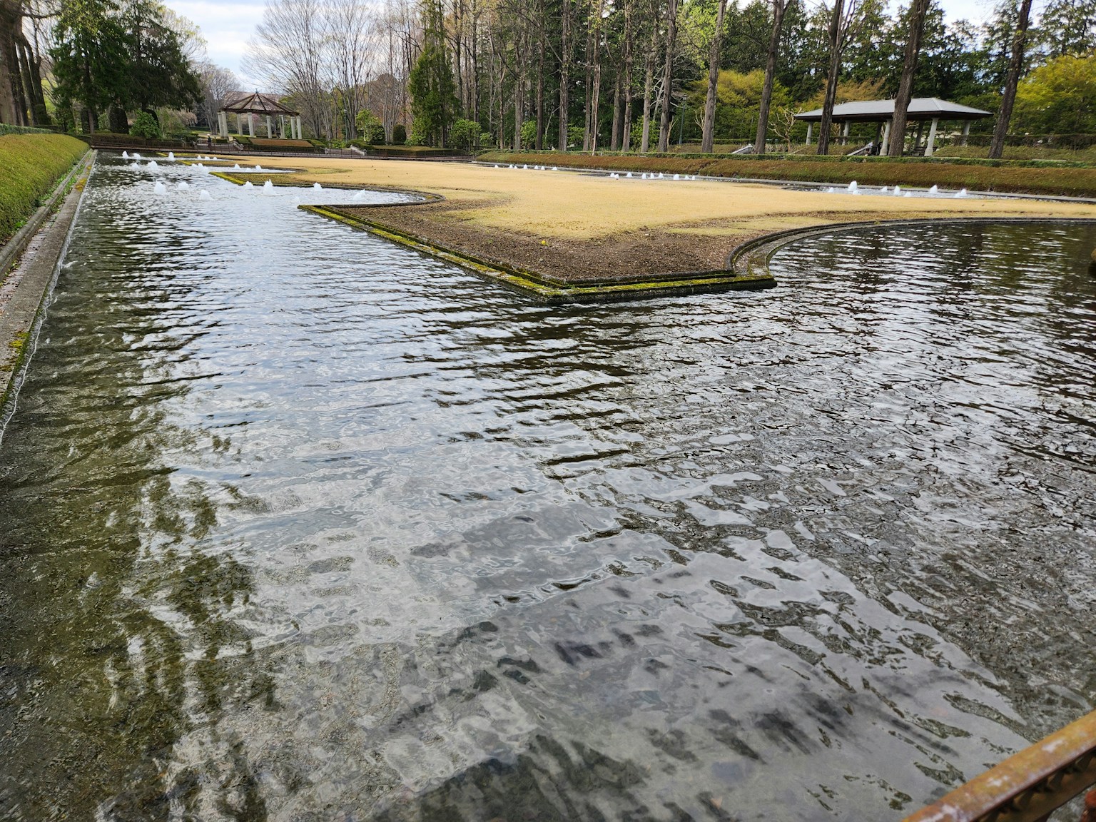 Kolam tenang dengan area pasir dikelilingi pepohonan rimbun dan gazebo