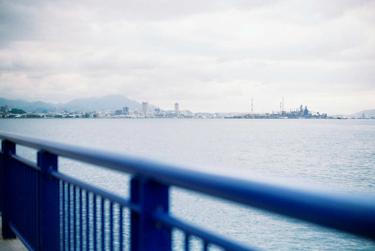 Una vista costera con una barandilla azul y un horizonte urbano distante bajo cielos nublados