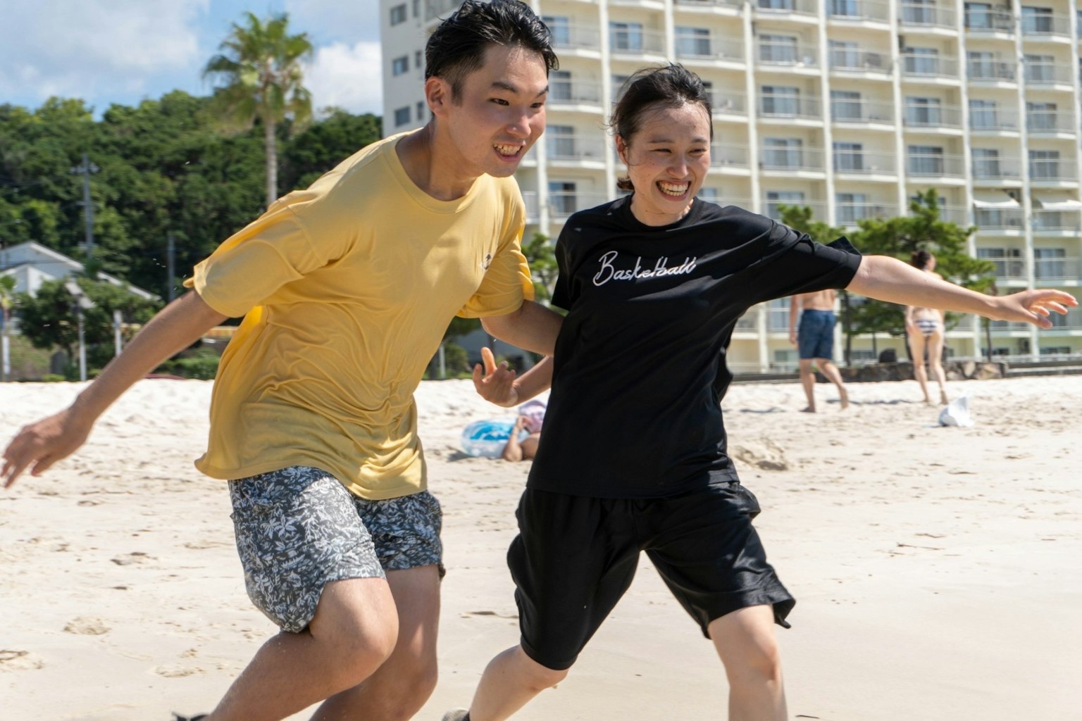 Zwei junge Menschen, die am Strand spielen, tragen bunte Kleidung und lächeln fröhlich, mit dem Ozean und Gebäuden im Hintergrund
