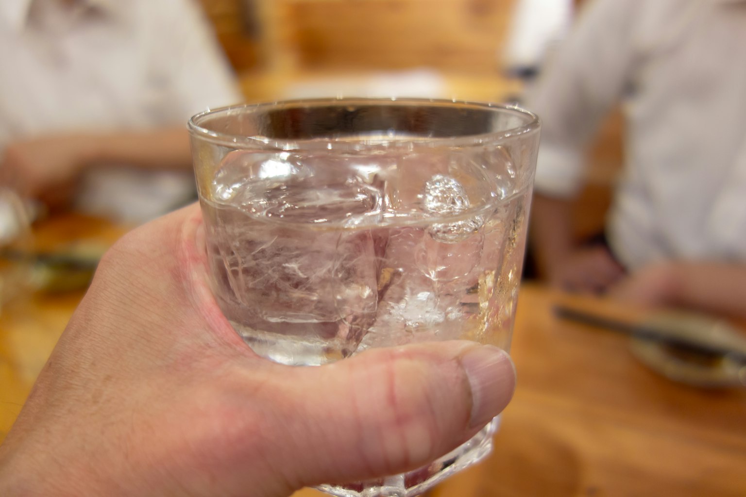 一只手握着冰水的玻璃杯，背景模糊的人物