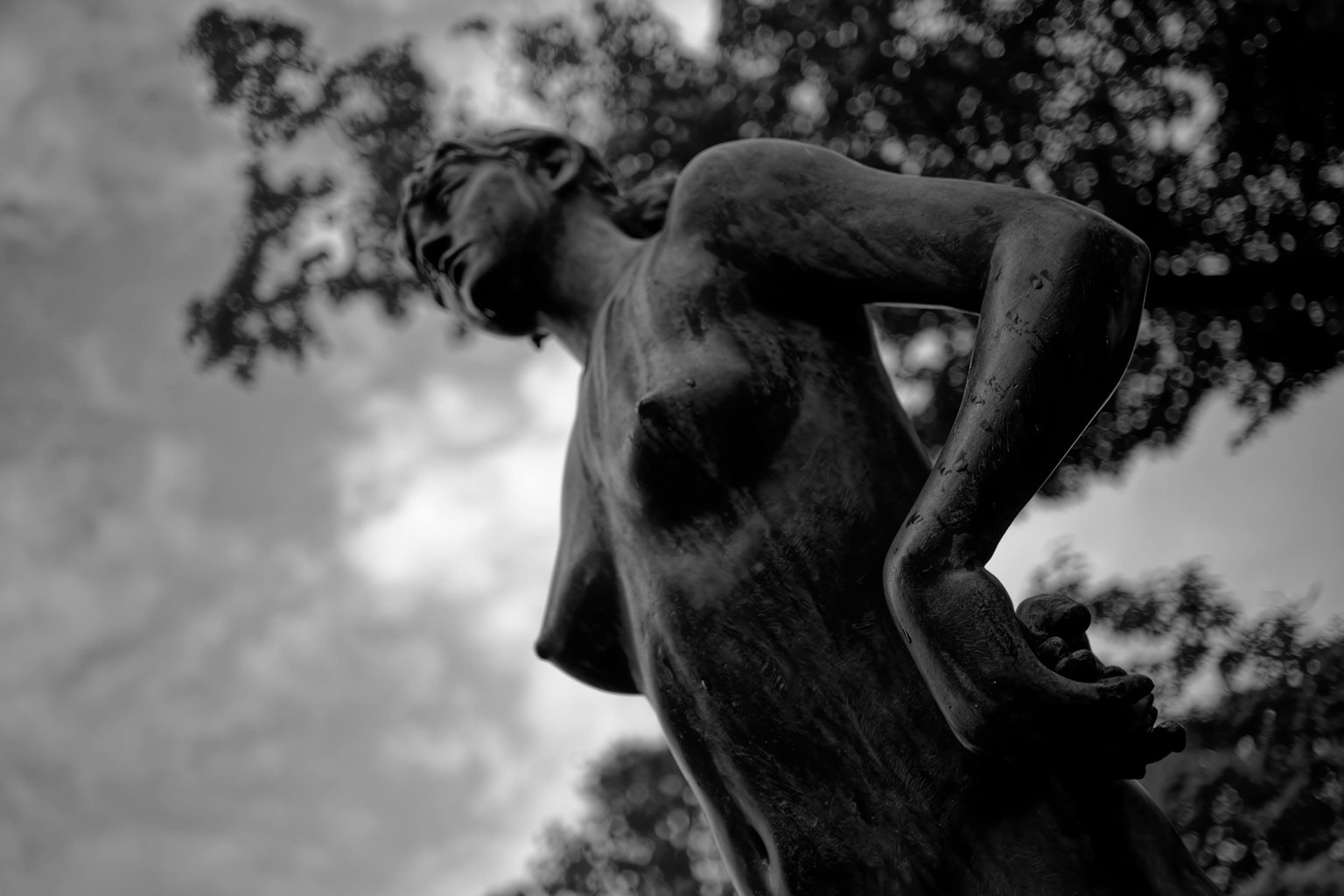 A monochrome photo of a female sculpture viewed from the side with trees and clouds in the background