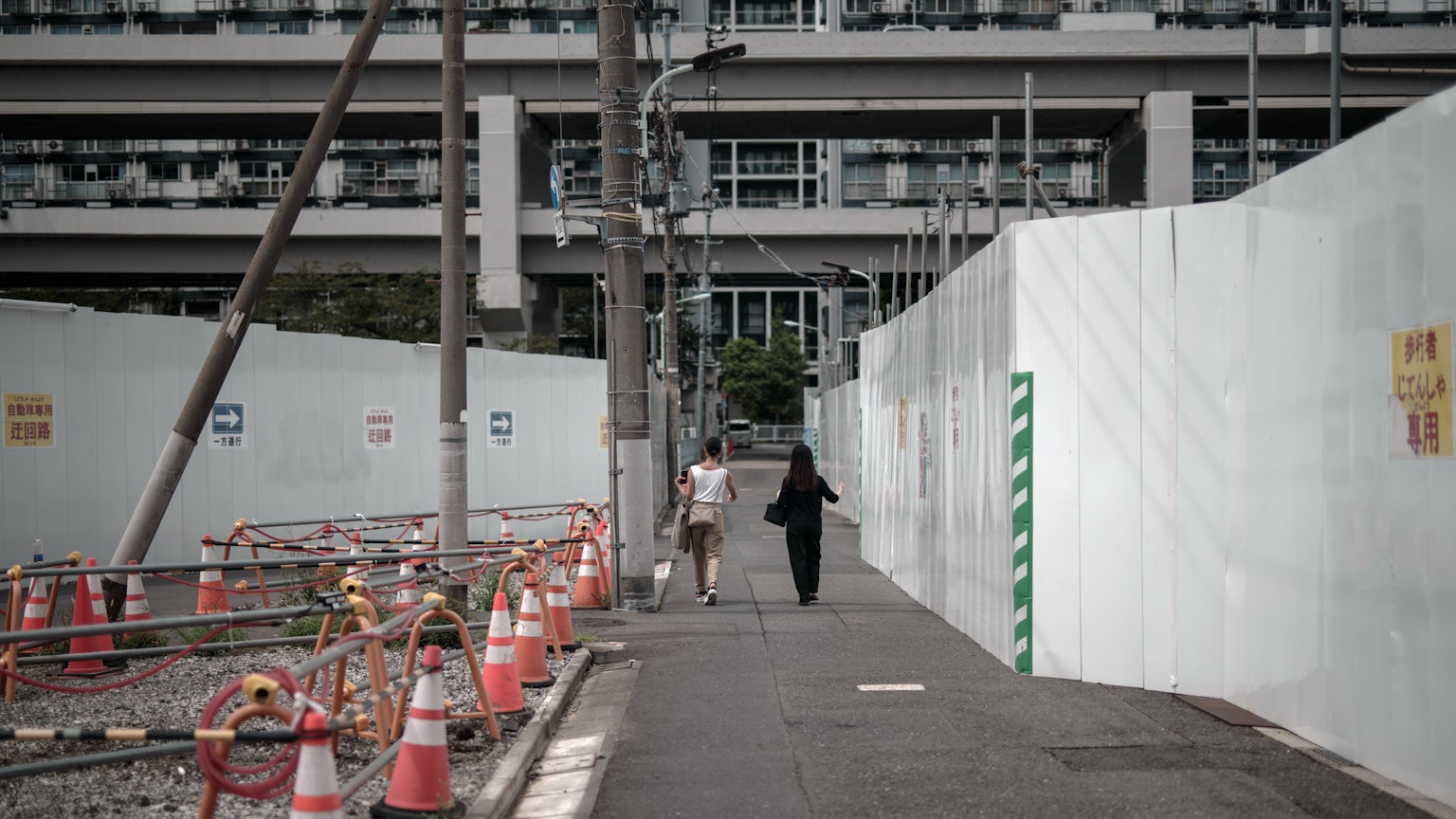 Two people walking on a street near a construction site White barriers and orange cones visible