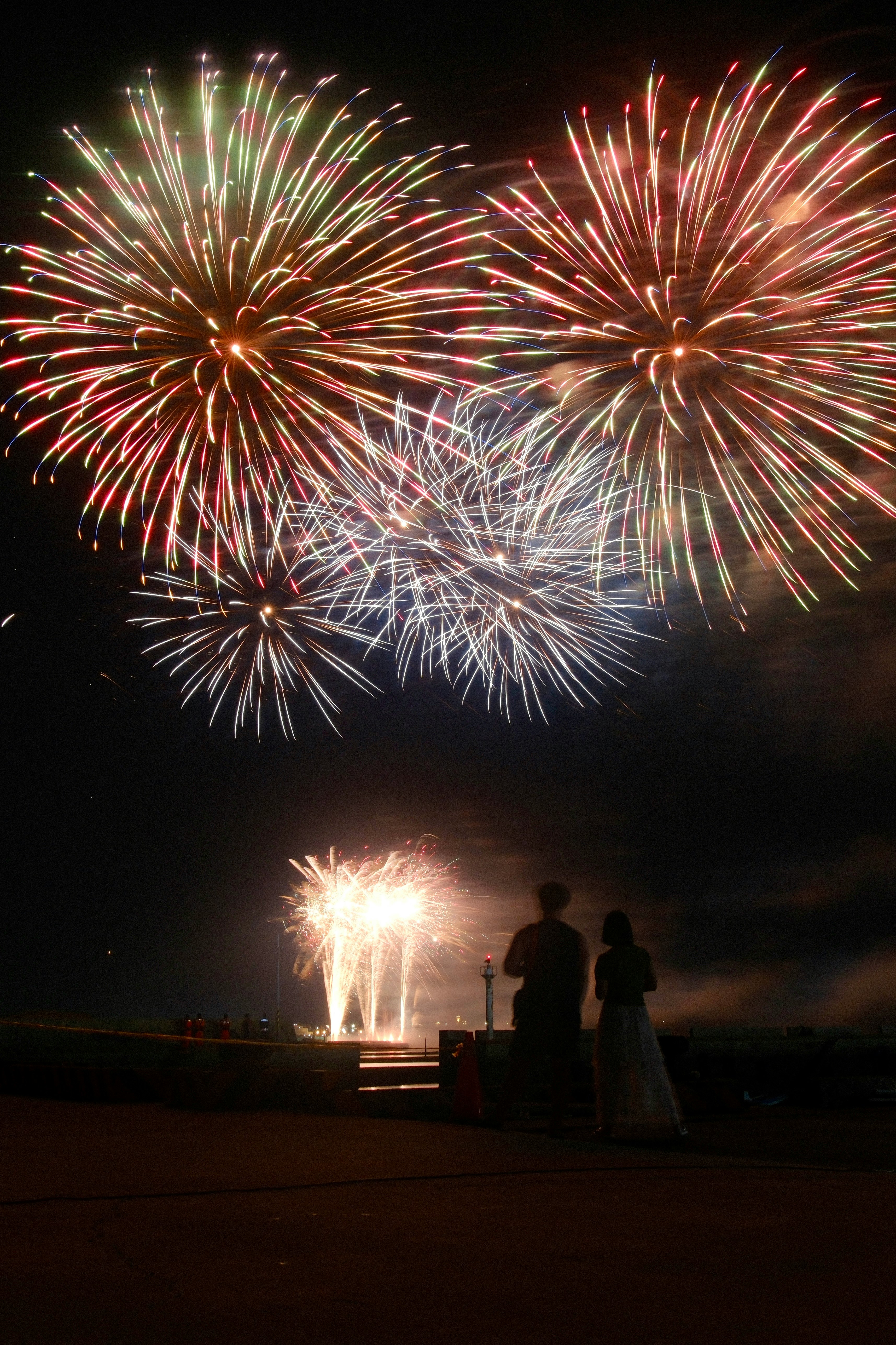 夜空に咲く花火と人影のある海辺の広場