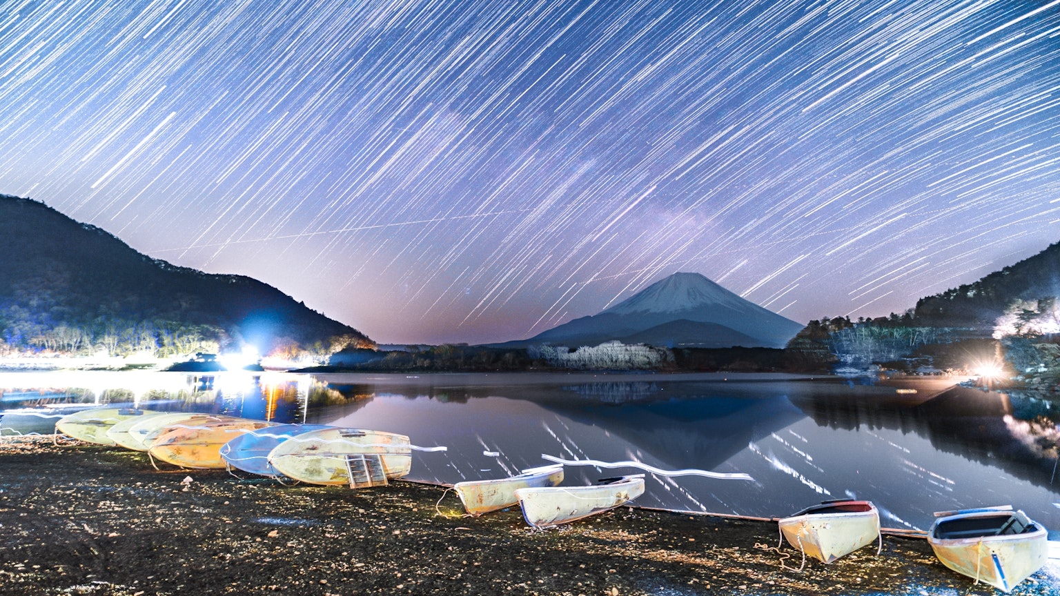 Star trails over Mount Fuji with a serene lake and boats in the foreground