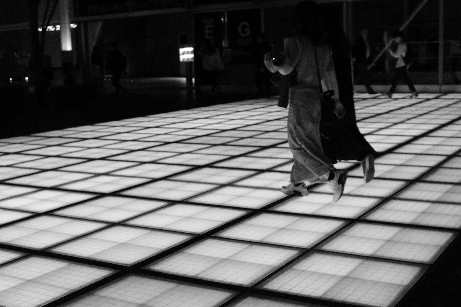Personas caminando sobre un piso en cuadrícula brillante en un entorno oscuro
