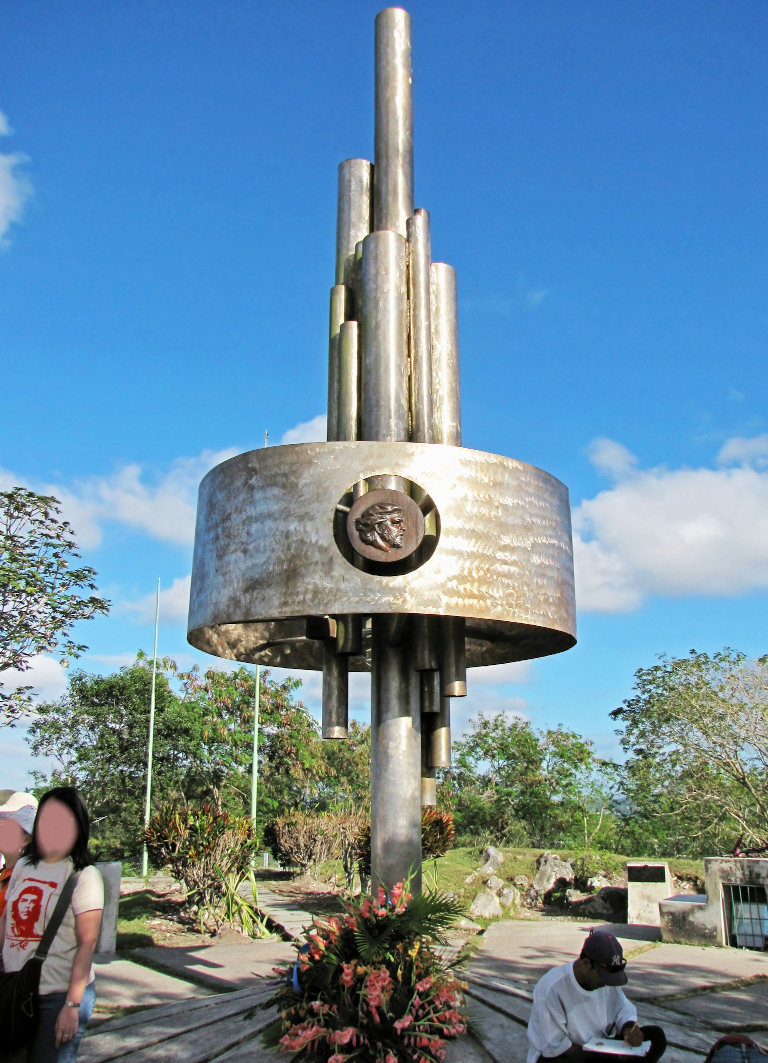 Monumen logam dengan persembahan bunga di bawah langit biru