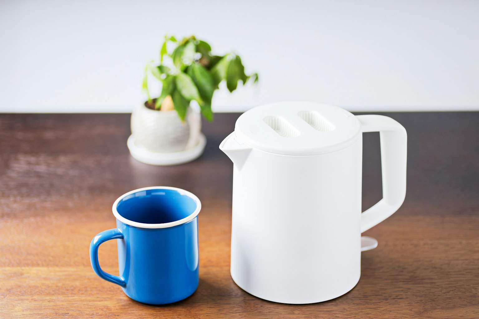 A white kettle and a blue mug placed on a wooden table with a small plant in the background