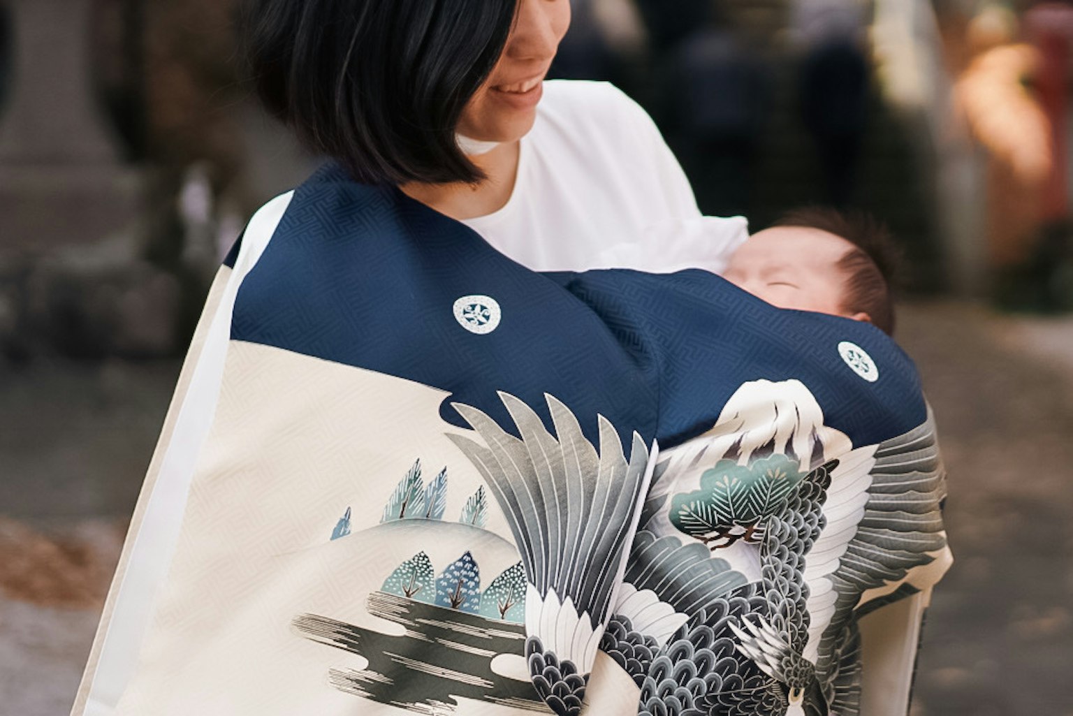 Una mujer sosteniendo a un bebé envuelto en una prenda tradicional con un diseño de grulla