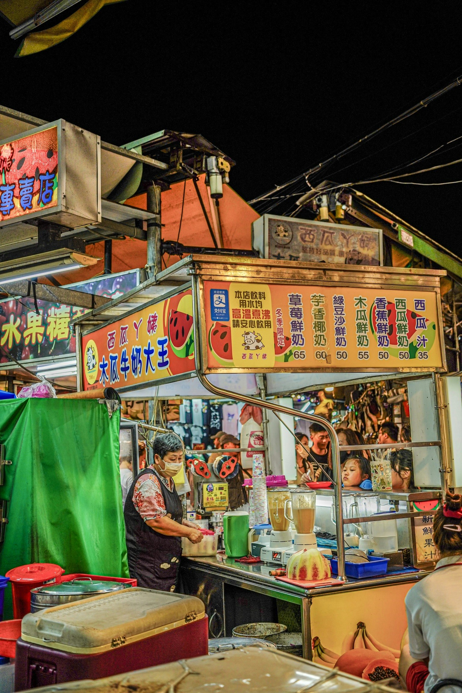 Ein Verkäufer, der Getränke an einem Nachtmarkt mit bunten Schildern und lebhafter Atmosphäre zubereitet
